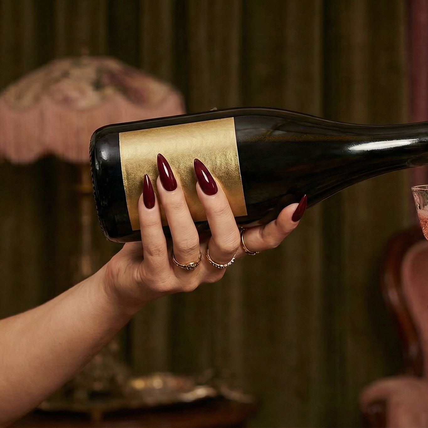 A woman's hand with dark red nails and multiple rings holding a champagne bottle with gold labeling, ready to pour.