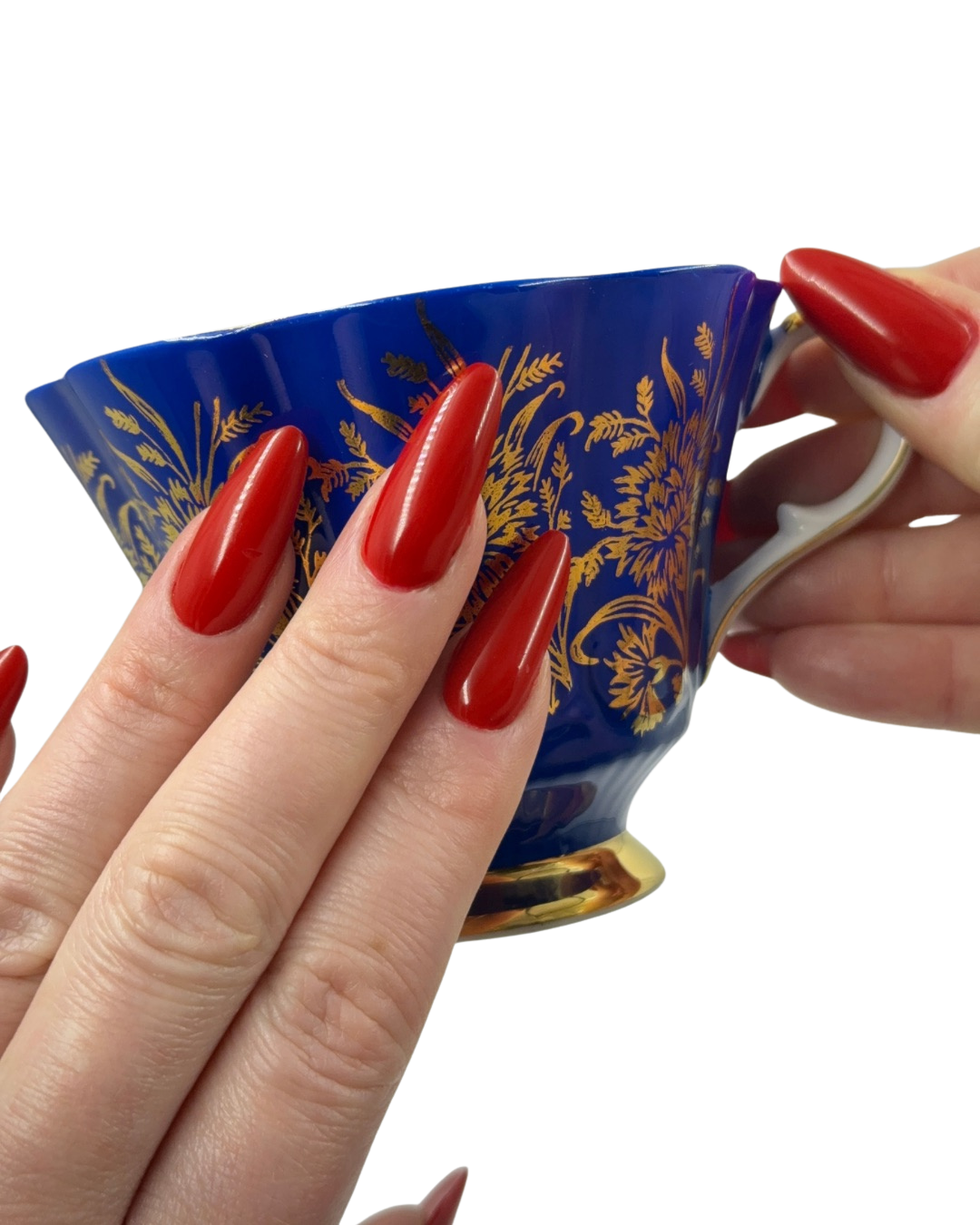 Close-up of a person's hand with long, shiny red gel nails holding a decorative blue and gold teacup.