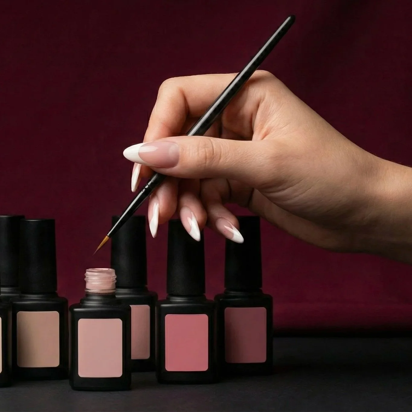 A hand with long nails holds a fine-tipped brush above a row of bottles containing various shades of pink nail polish, set against a dark red background.