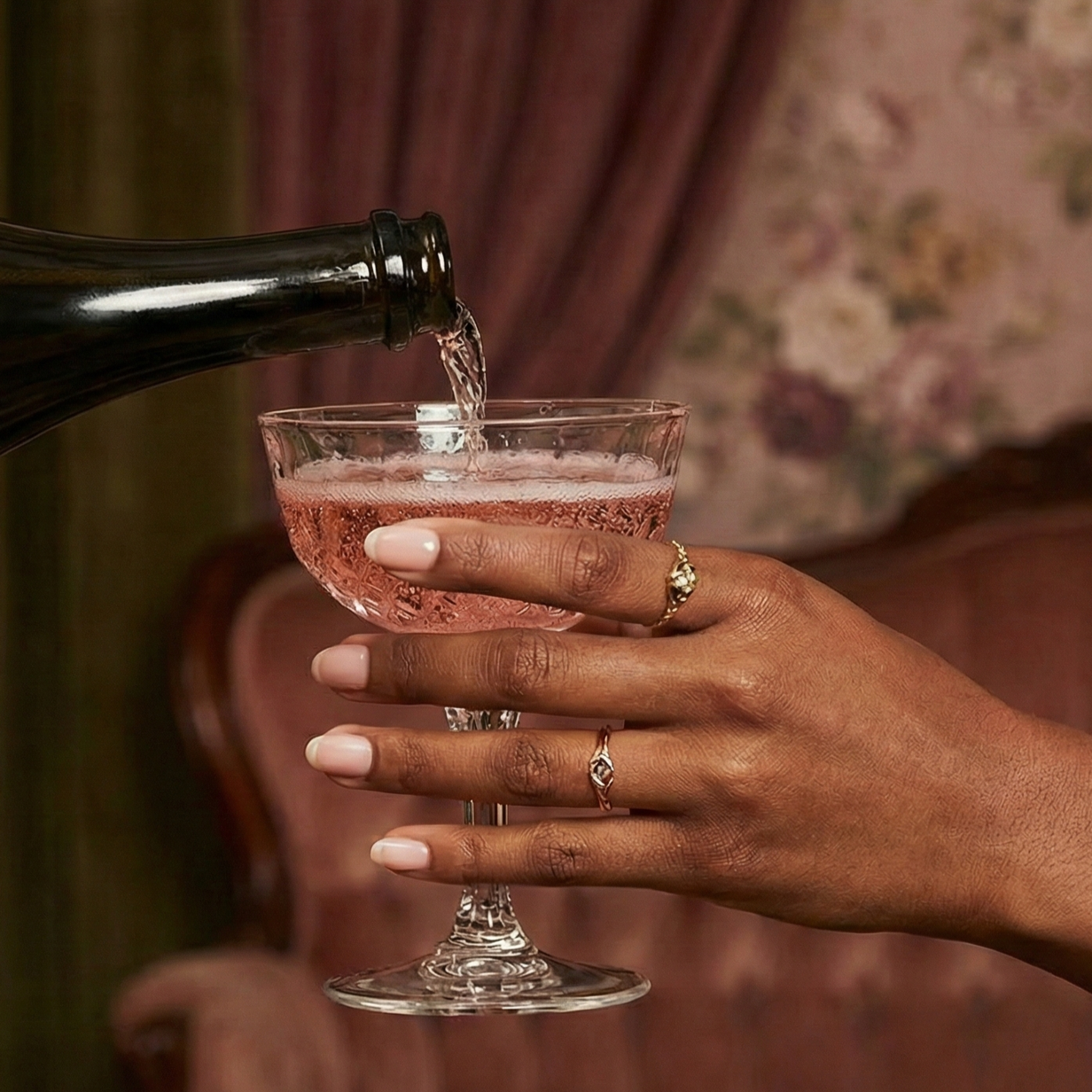 A person with dark skin wearing rings on their fingers holds a vintage-style cocktail glass filled with a pink beverage, while someone pours a drink into it from a dark bottle. The background features soft, floral-patterned fabric.