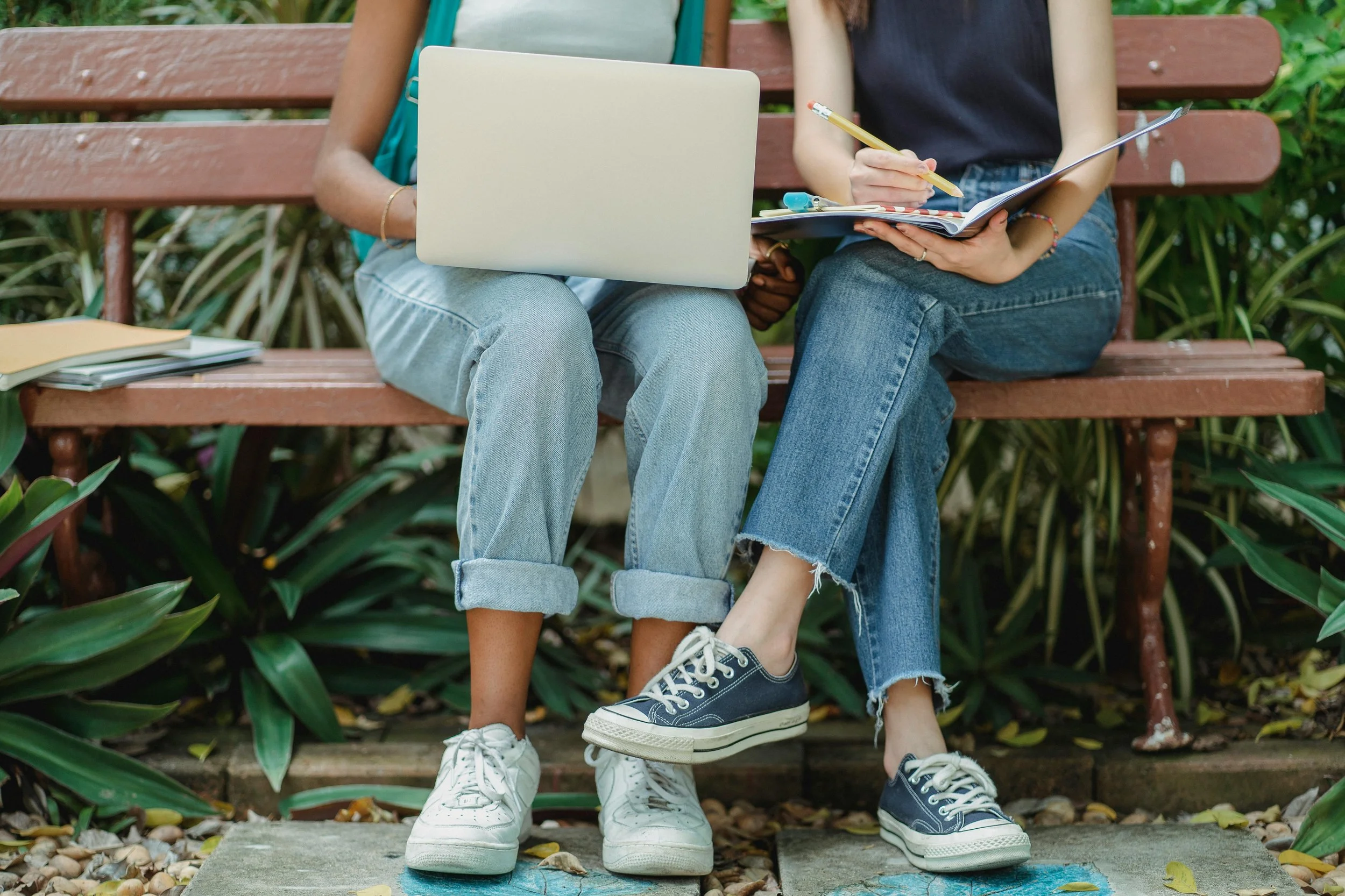 Two people sitting on a park bench with notebooks and a laptop.