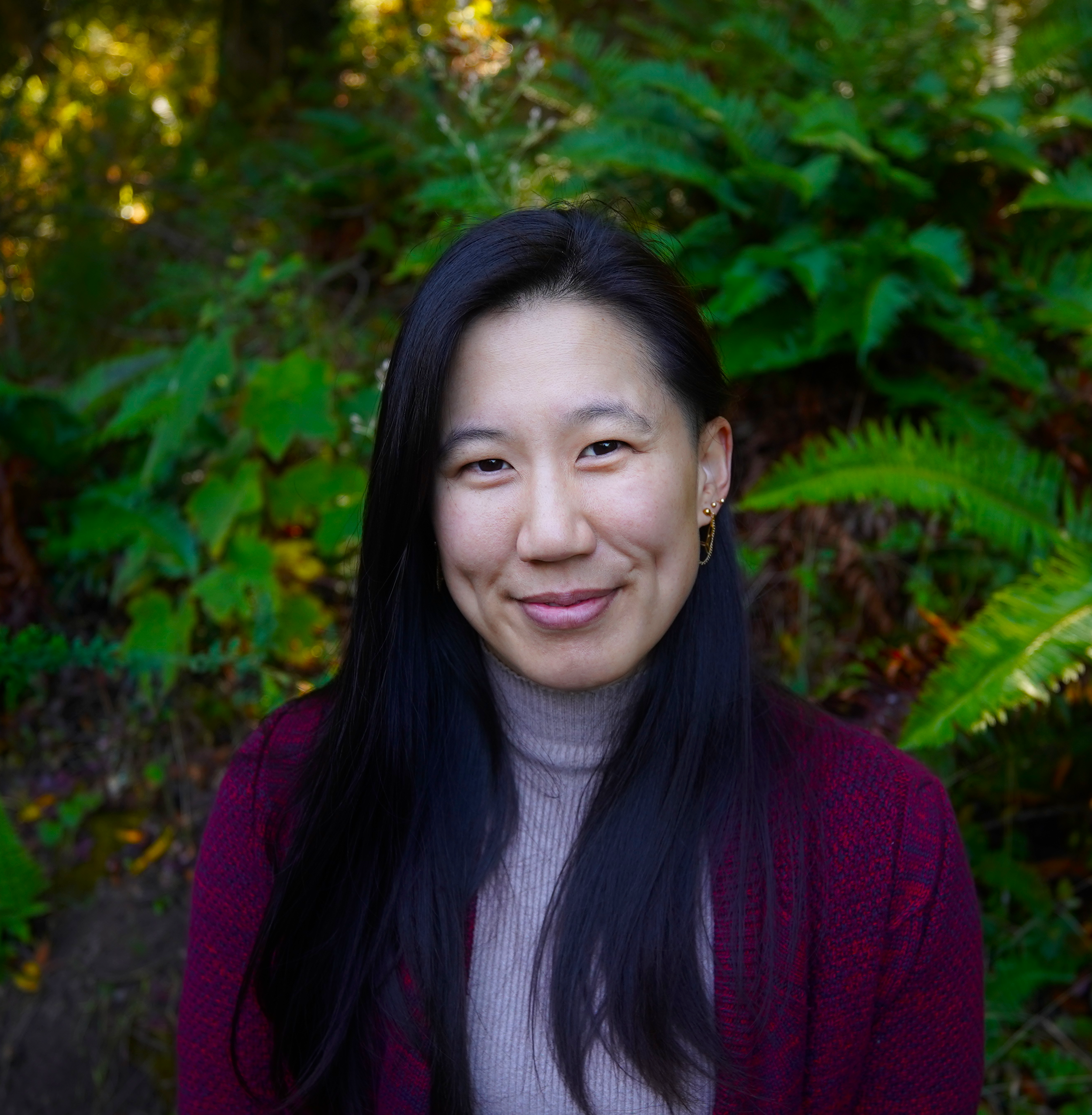 A woman with long black hair smiling outdoors with green foliage in the background.