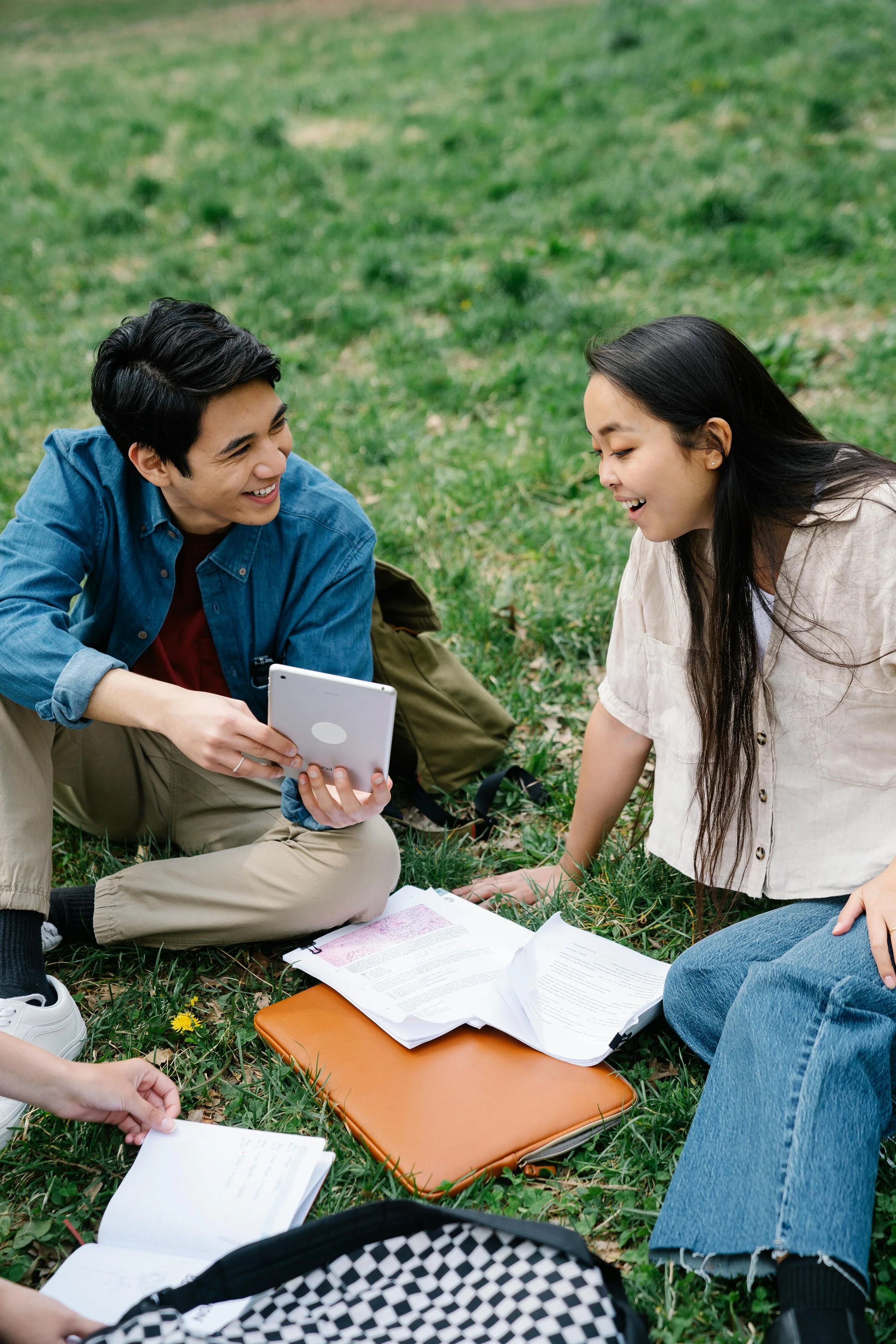 Two young people, a man and a woman, sitting on the grass outdoors, smiling, with textbooks, notebooks, and a tablet, engaged in studying or conversation.