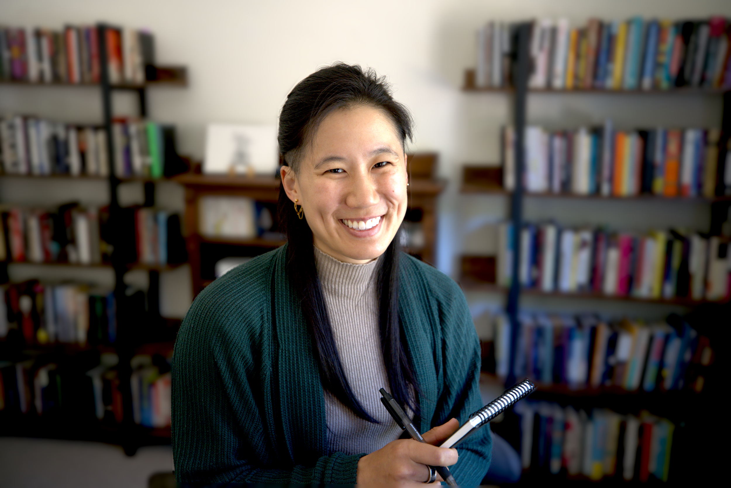 A woman smiling while holding a pen and notebook, sitting in front of bookshelves filled with books.