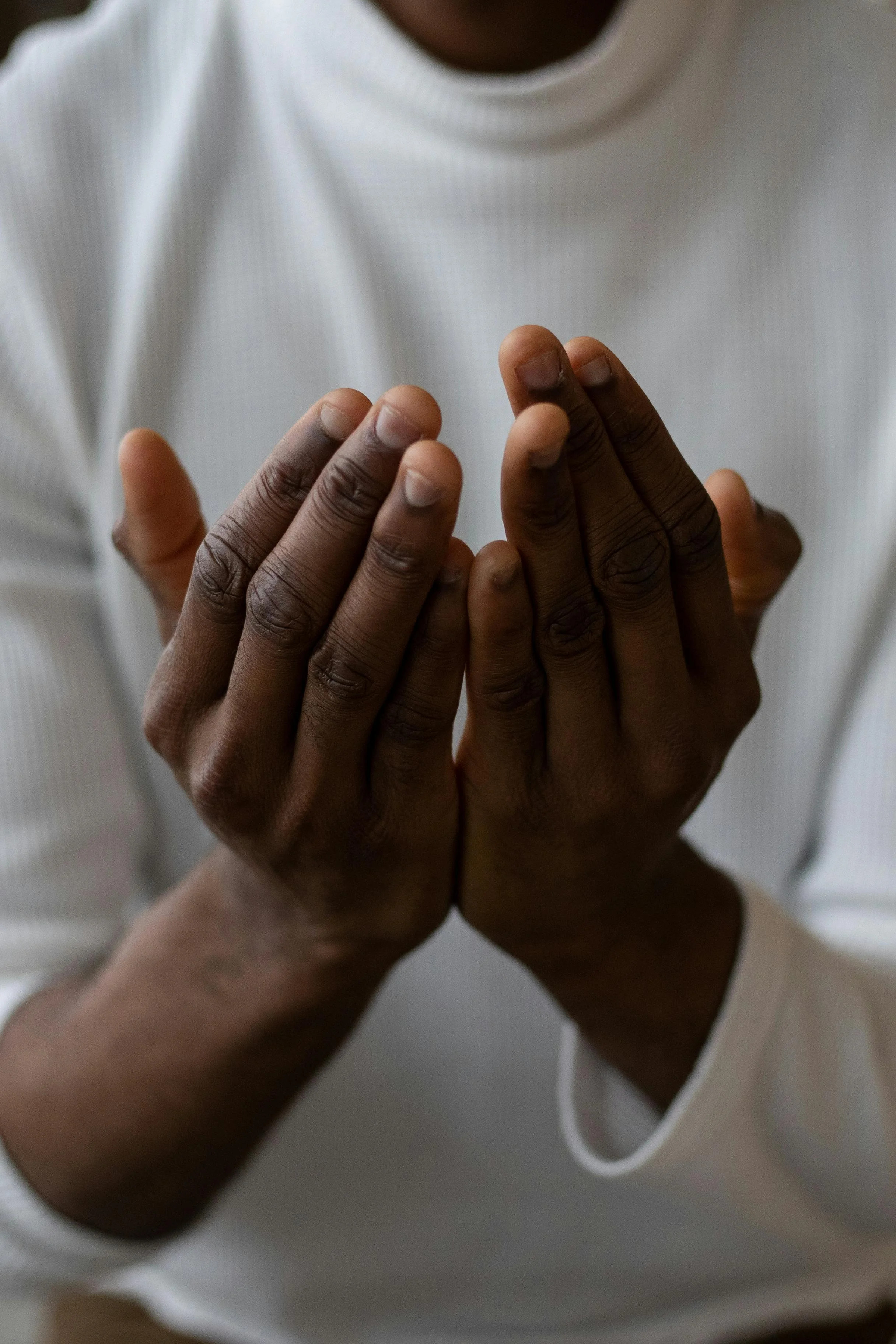 A person with dark skin wearing a white long-sleeve shirt holding their hands together in a praying position.