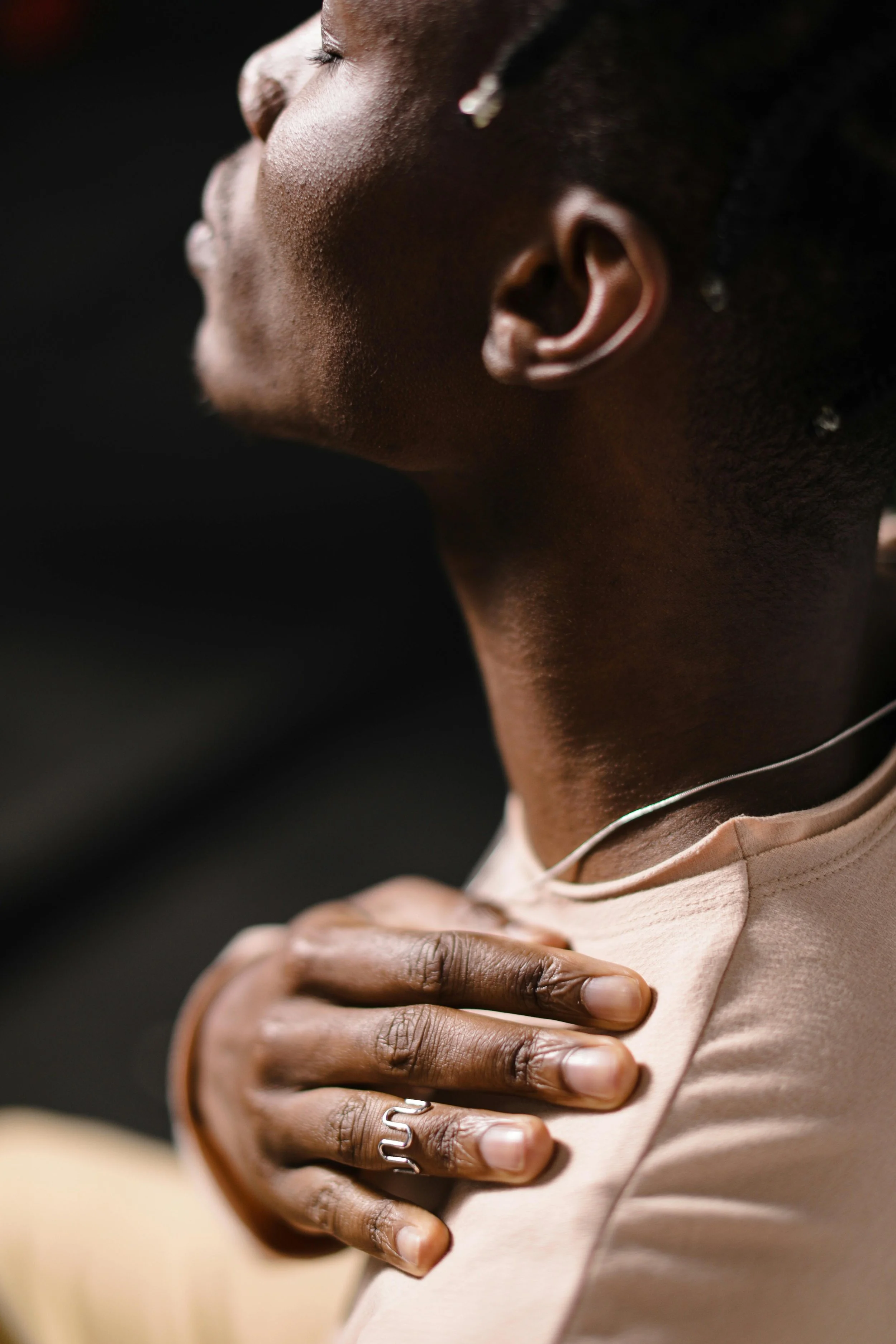 Close-up of a person with dark skin touching their chest with their hand, wearing a ring and a beige shirt. Their face is partially visible, showing a side profile with closed eyes and earrings, against a dark background.