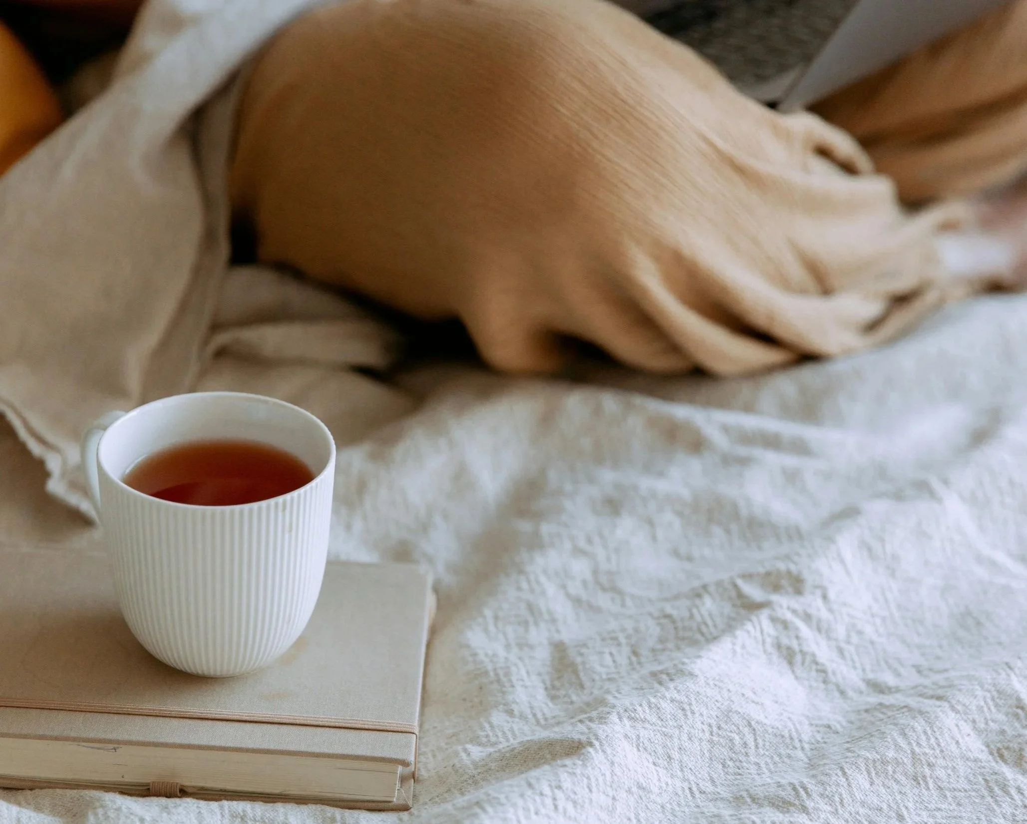 A white mug filled with tea resting on top of a closed beige notebook, placed on a soft white textured fabric surface with a beige blanket and a pillow in the background.