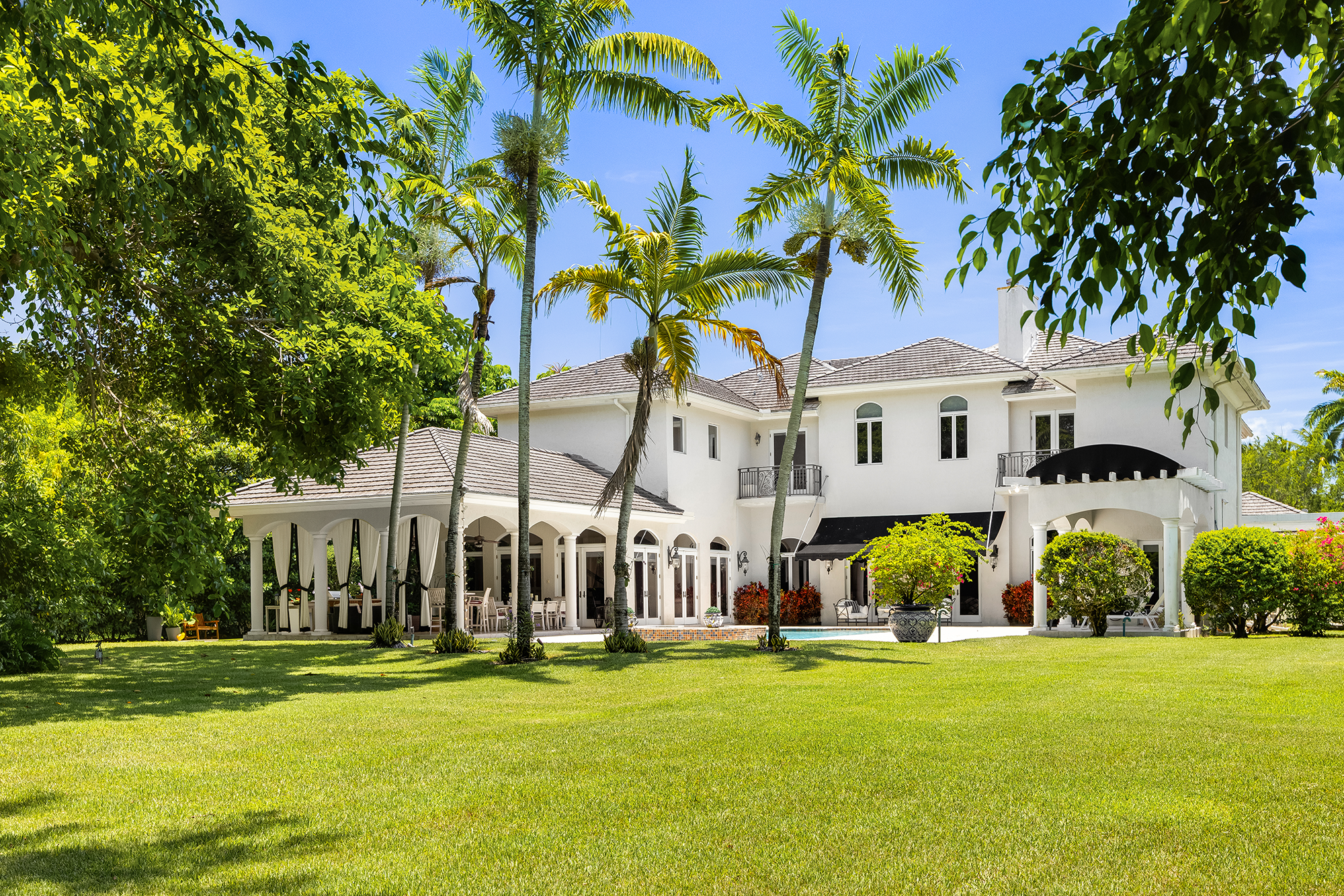 A large two-story white house with a tiled roof, surrounded by a well-maintained grassy yard with tropical trees including palm trees, under a bright blue sky.