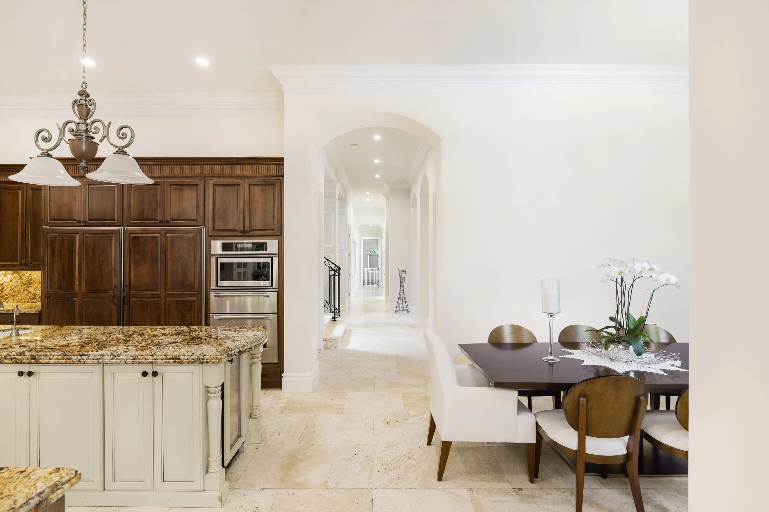 View of a kitchen and dining area with white walls, beige tile flooring, and wooden cabinetry. The kitchen features a granite countertop and built-in stainless steel appliances. The dining table has a flower arrangement with white orchids and candles
