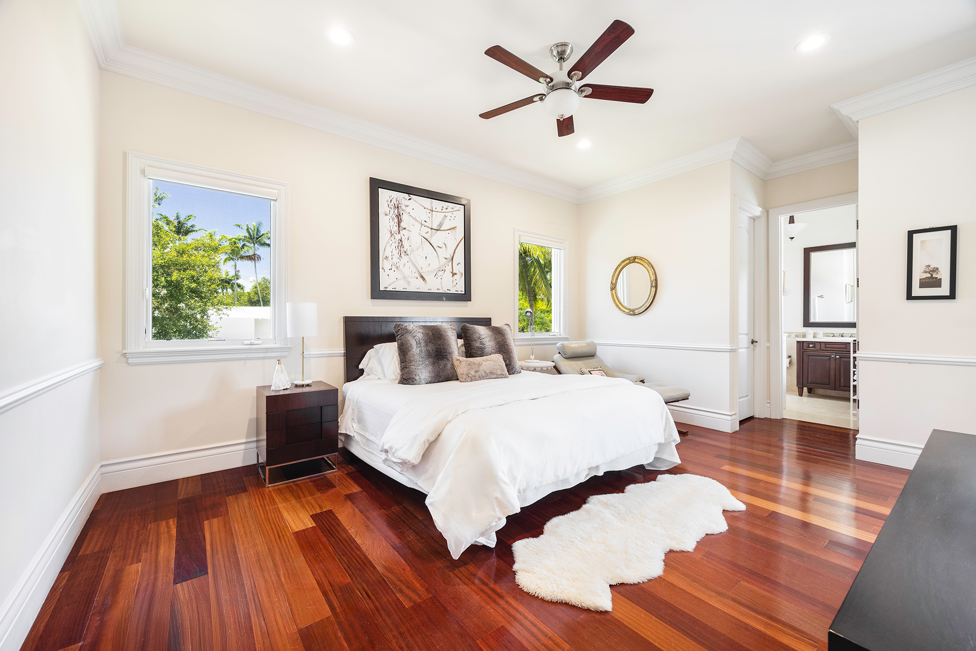 Bright bedroom with white walls, hardwood floors, a ceiling fan, a large bed with dark headboard and pillows, two windows showing green trees outside, a nightstand with a lamp, and a bathroom visible through an open door.