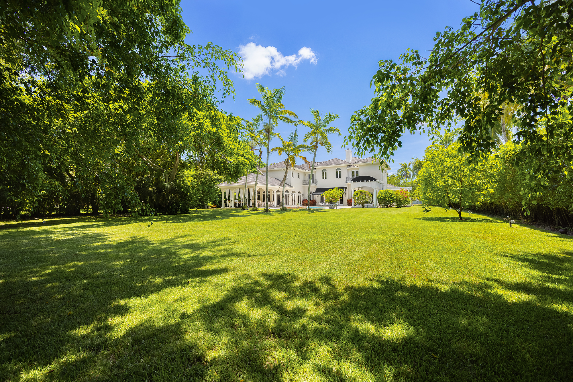 Large white house with multiple floors and arches, surrounded by lush green trees and a well-maintained grassy lawn, under a bright blue sky with a few clouds.