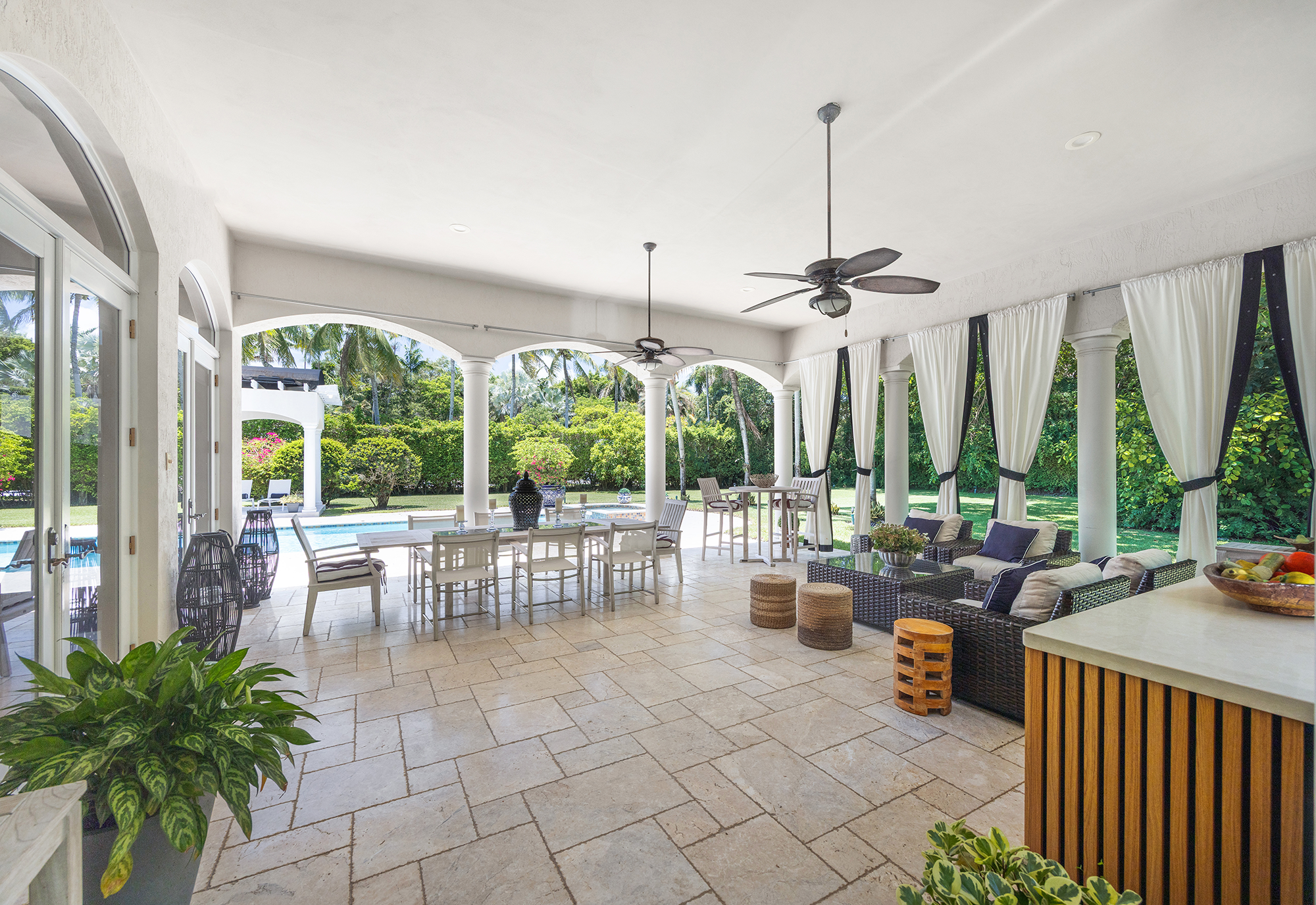 Covered patio with ceiling fans, white curtains, outdoor furniture, and a view of a backyard with a pool, palm trees, and greenery.