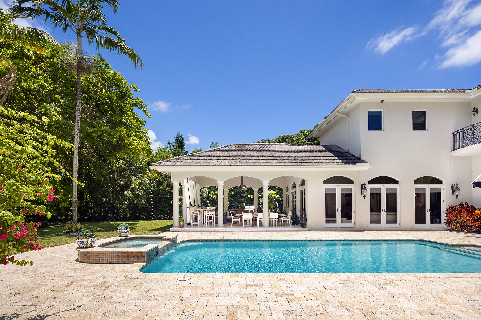 Backyard with swimming pool, hot tub, patio table and chairs, white house with large glass doors, lush green trees, and clear blue sky.