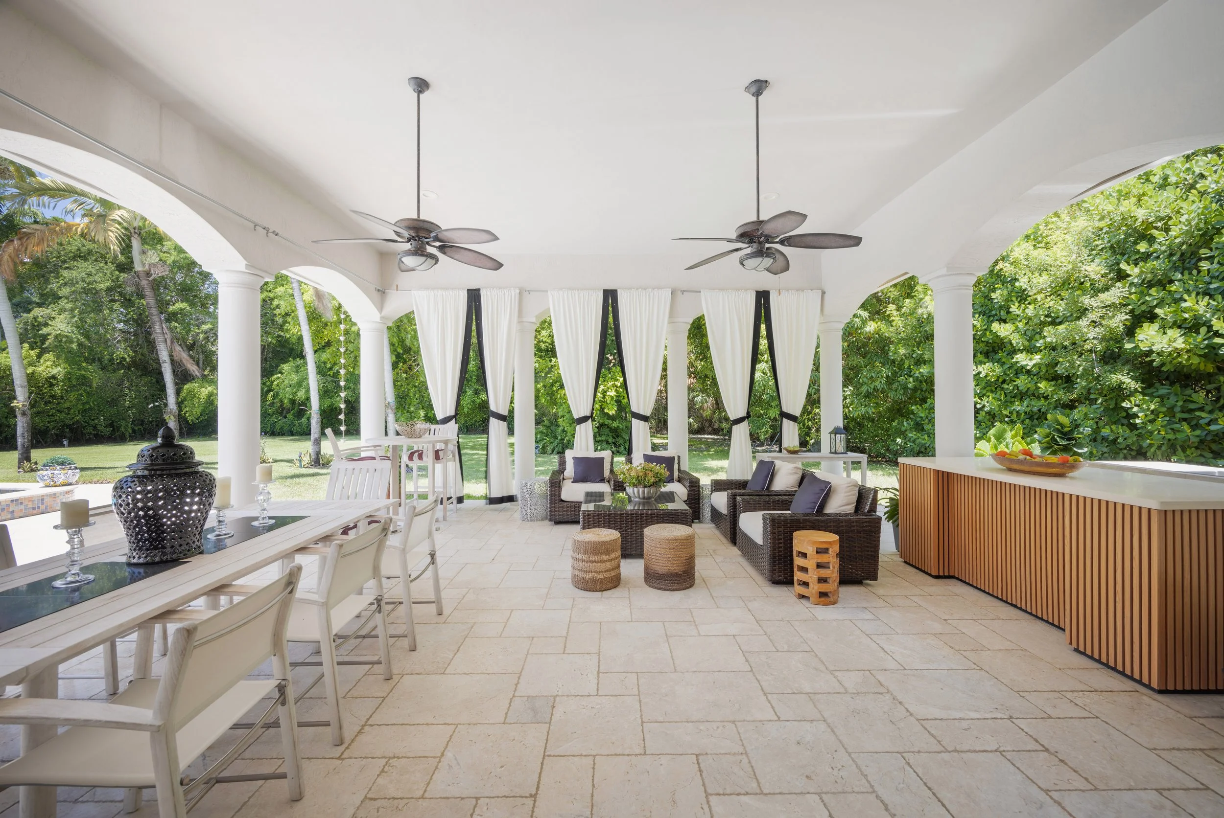 Spacious outdoor patio with white ceiling, ceiling fans, black and white curtains, wicker seating with cushions, a long dining table with chairs, decorative jars, and views of lush green trees.