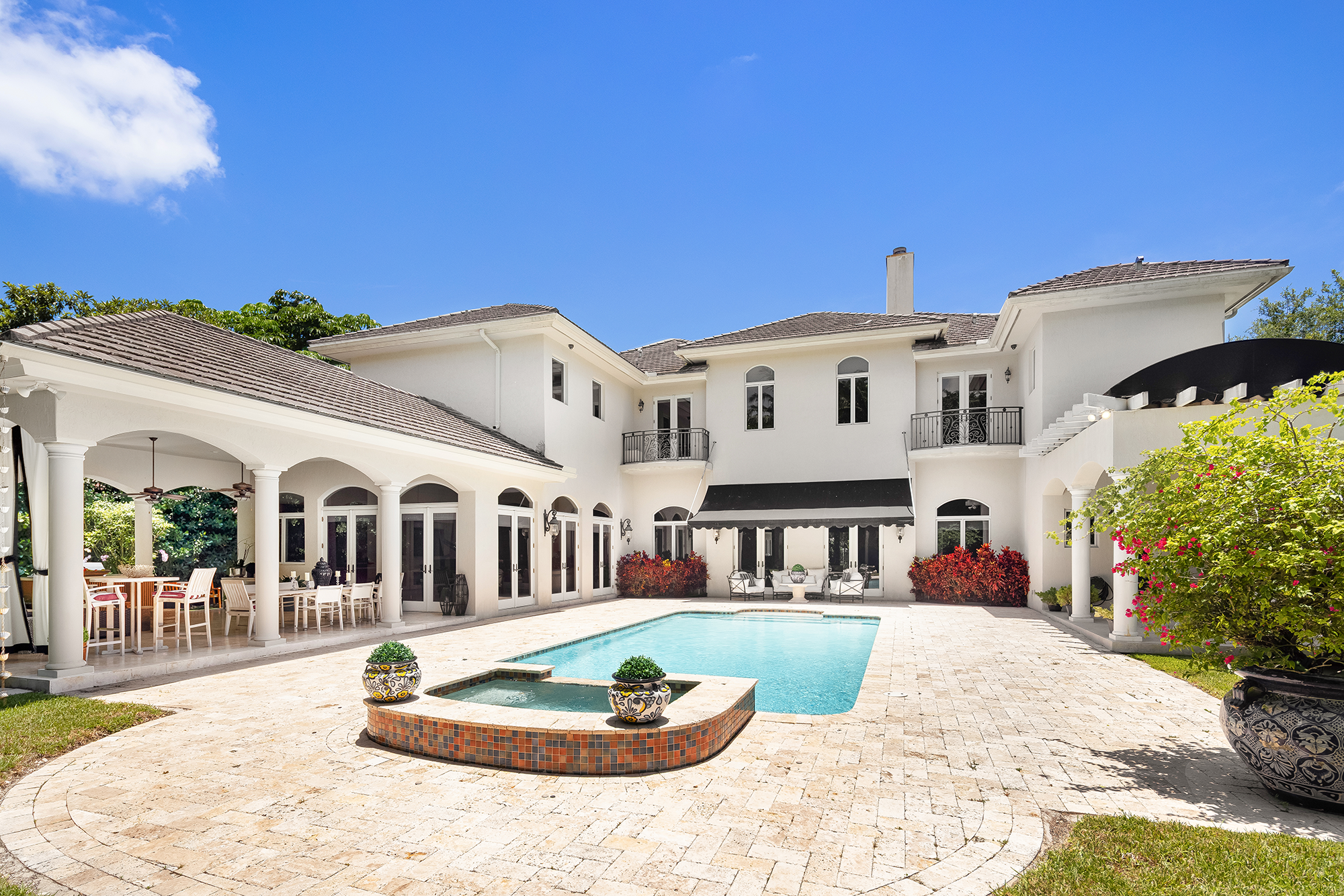 Large white house with multiple levels, arched windows, and balconies, surrounded by a backyard with a pool, patio furniture, and potted plants, under a blue sky.