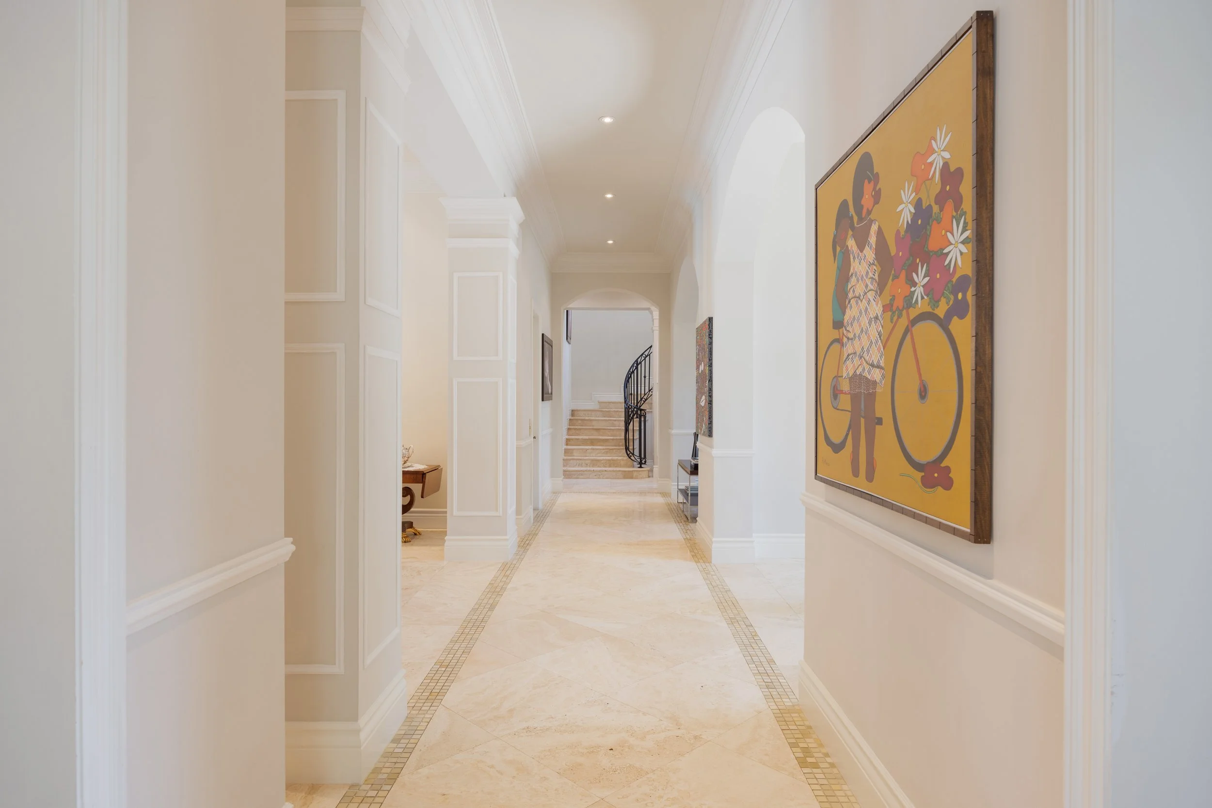Elegant hallway with beige marble floors, white walls, framed artwork, and a staircase at the end.