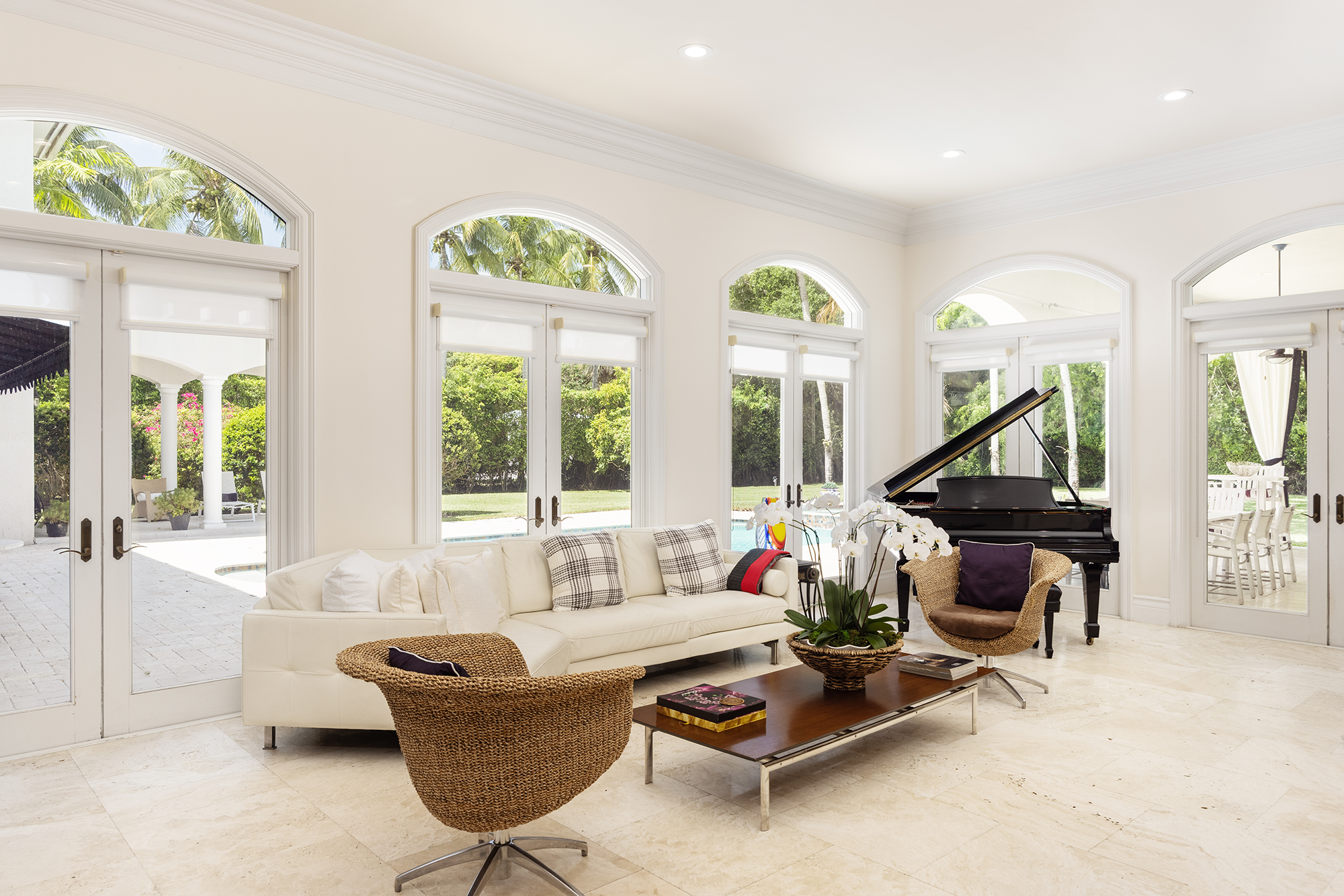 Bright living room with white walls, large windows with white blinds, and a view of a tropical outdoor patio and garden. Features include a white sofa with cushions, two woven chairs, a wooden coffee table with books and a plant, and a black grand pi