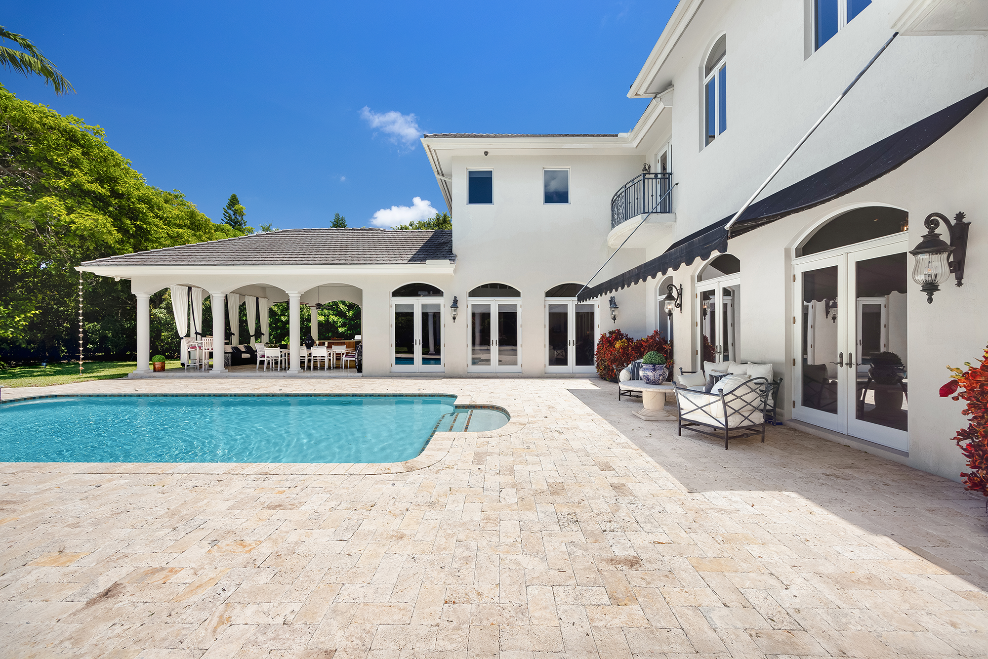 Backyard view of a white two-story house with a swimming pool, outdoor patio, and seating area under a black awning, surrounded by green trees and a blue sky.