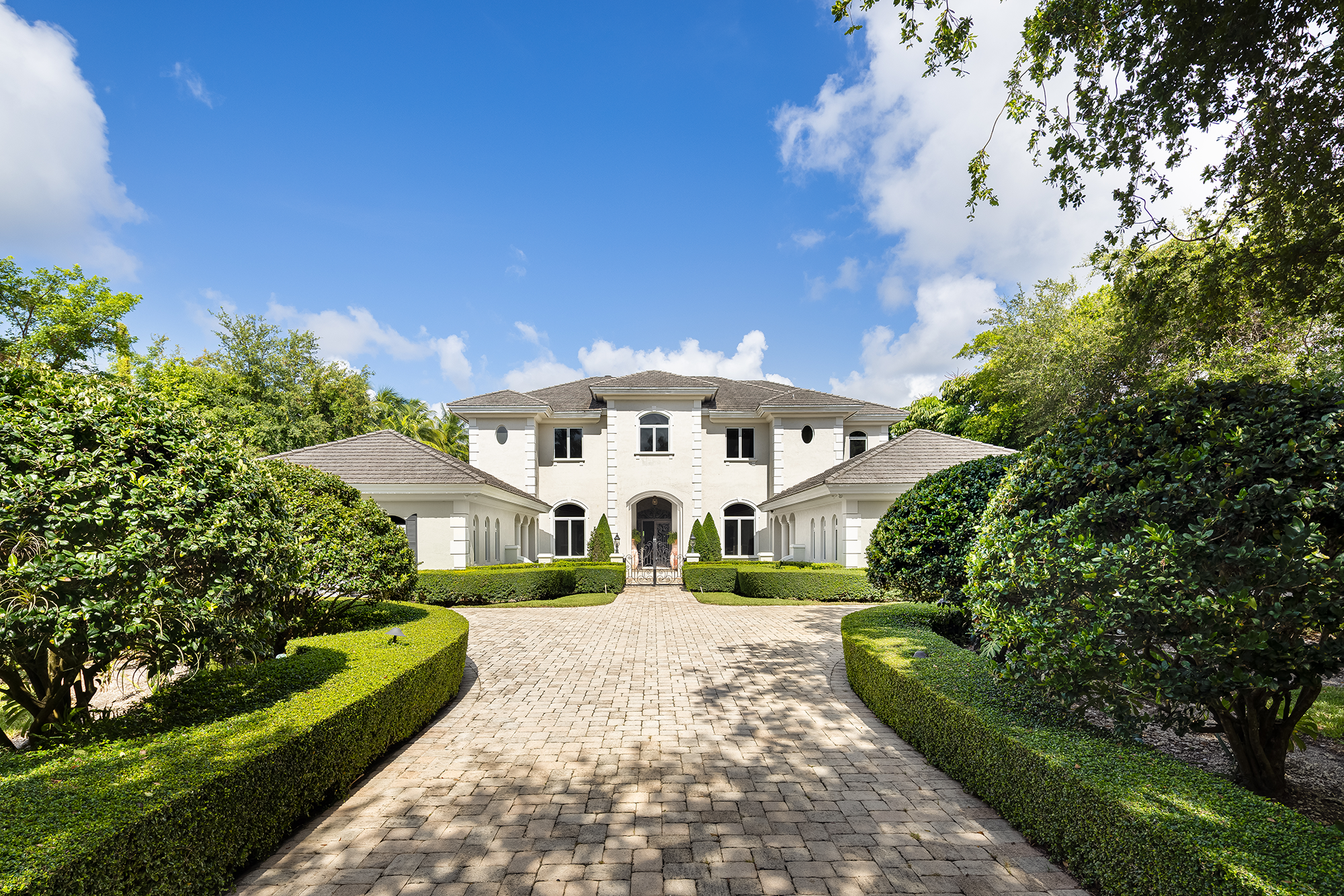 A large, white house with multiple windows and a central entrance, surrounded by a neatly landscaped garden with trimmed bushes and a brick pathway leading to the front door, under a partly cloudy blue sky.