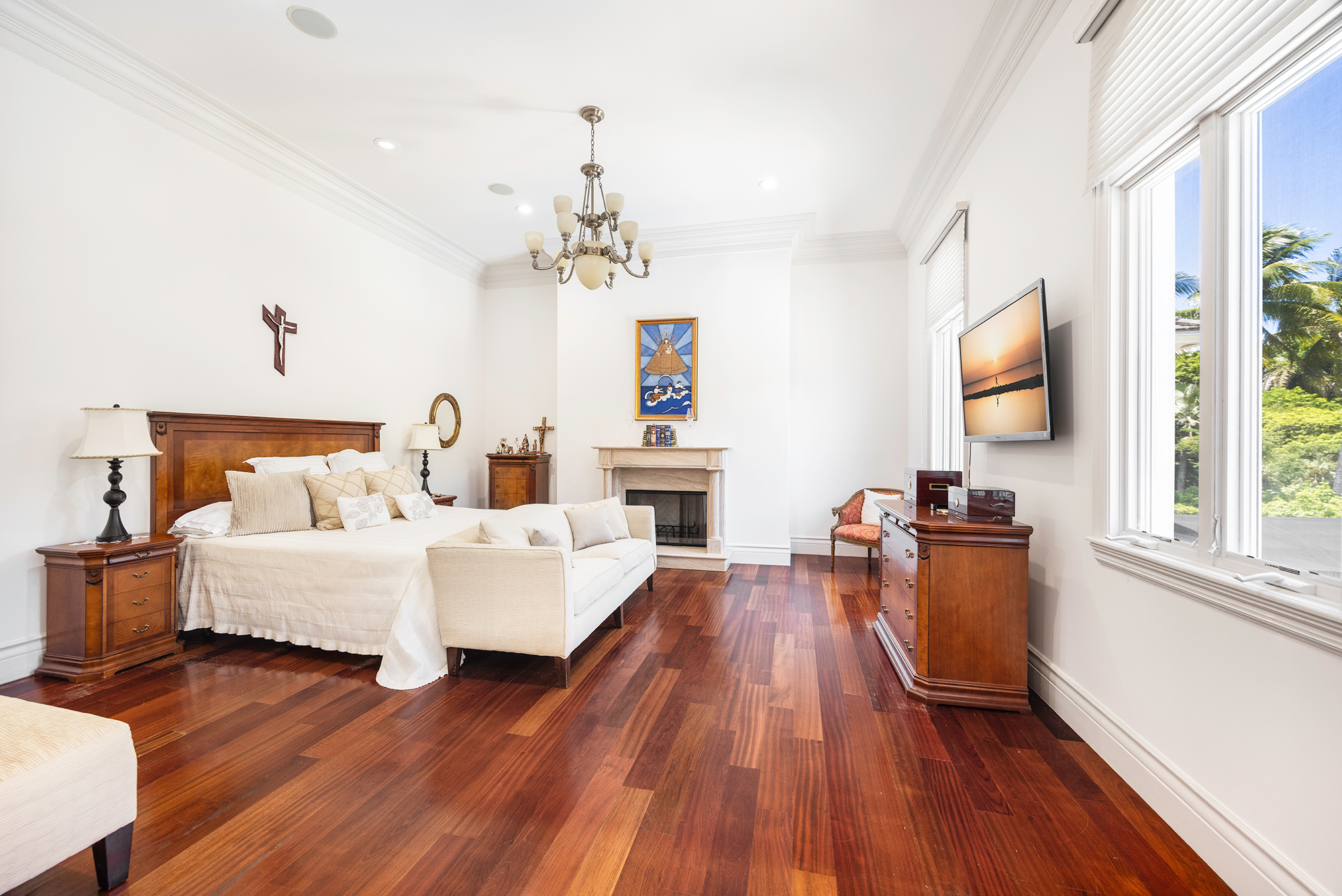 A spacious bedroom with white walls, hardwood floors, a large bed with beige bedding, a white sofa, wooden nightstands with lamps, a fireplace with religious artwork above, a wooden dresser, a wall-mounted TV, and large windows showing greenery outside.