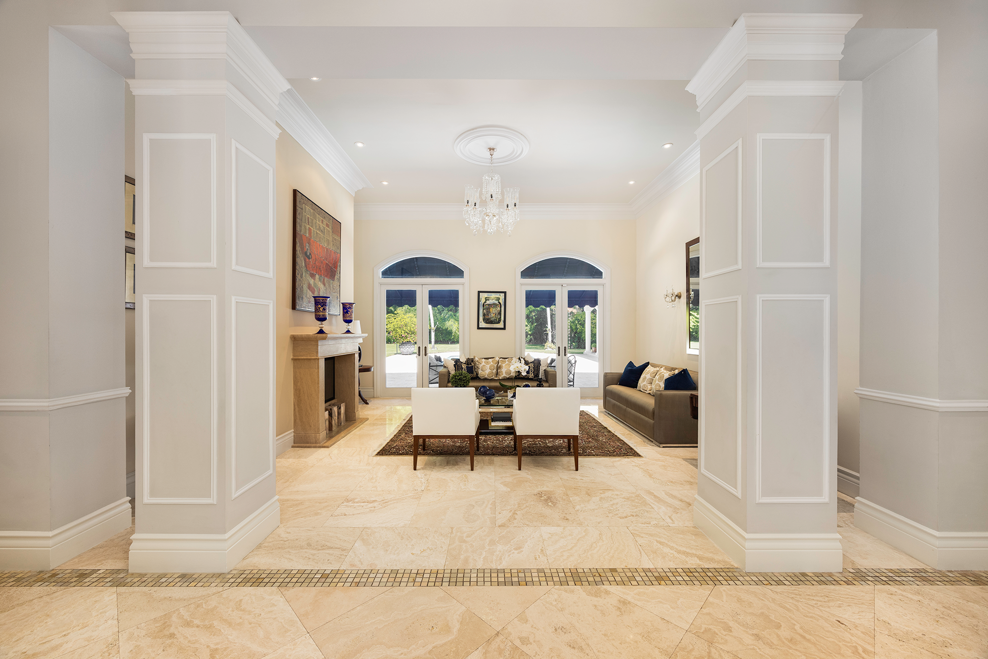 Bright and spacious living room with white walls, high ceilings, elegant chandelier, large arched windows, and various seating arrangements including a sofa and armchairs.