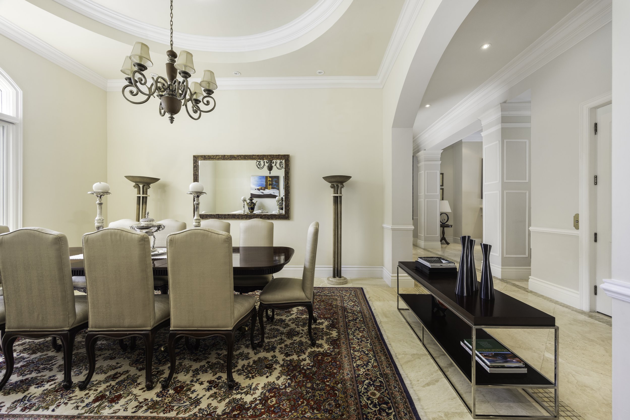 Elegant dining room with round wooden table surrounded by beige upholstered chairs, chandelier hanging above, decorative rugs, large mirror on cream-colored wall, and modern black console table with vases, in a spacious home interior.