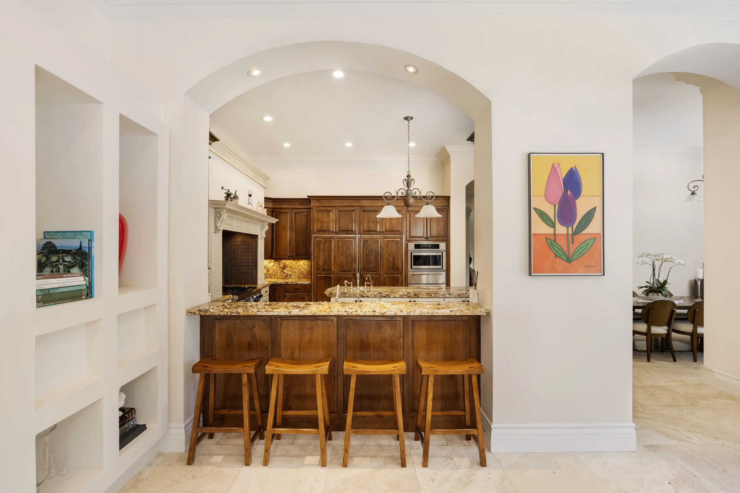 Kitchen with wooden cabinets, granite countertops, and a breakfast bar with four wooden stools. There's a built-in oven, a chandelier, and a decorative tile backsplash. A white wall with a colorful tulip artwork is on the right, and part of a dining area with chairs and a table is visible in the background.
