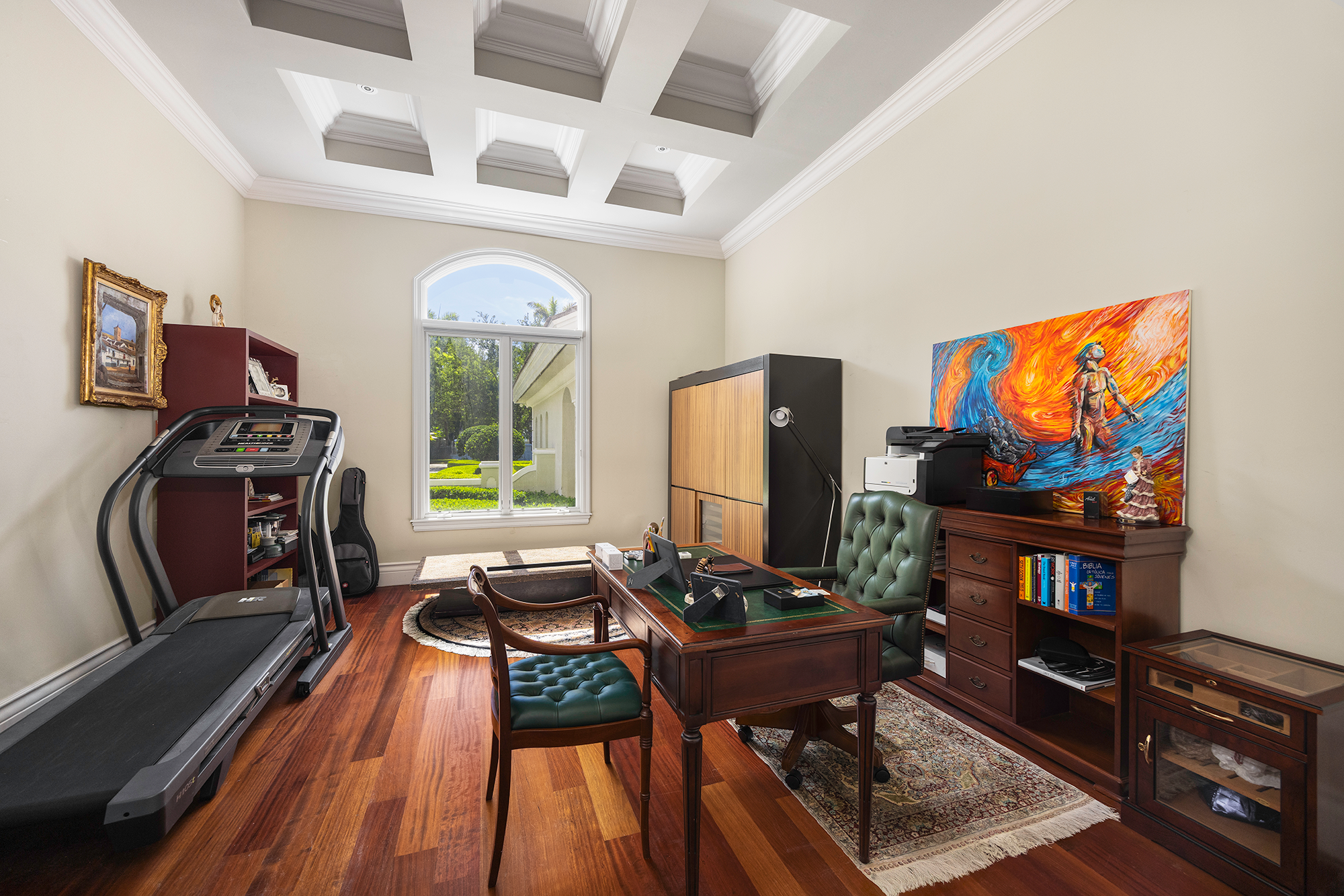 Home office with wooden desk, green tufted office chair, treadmill on left, window with greenery outside, abstract artwork with bright colors on right, bookshelves, and a wooden cabinet.