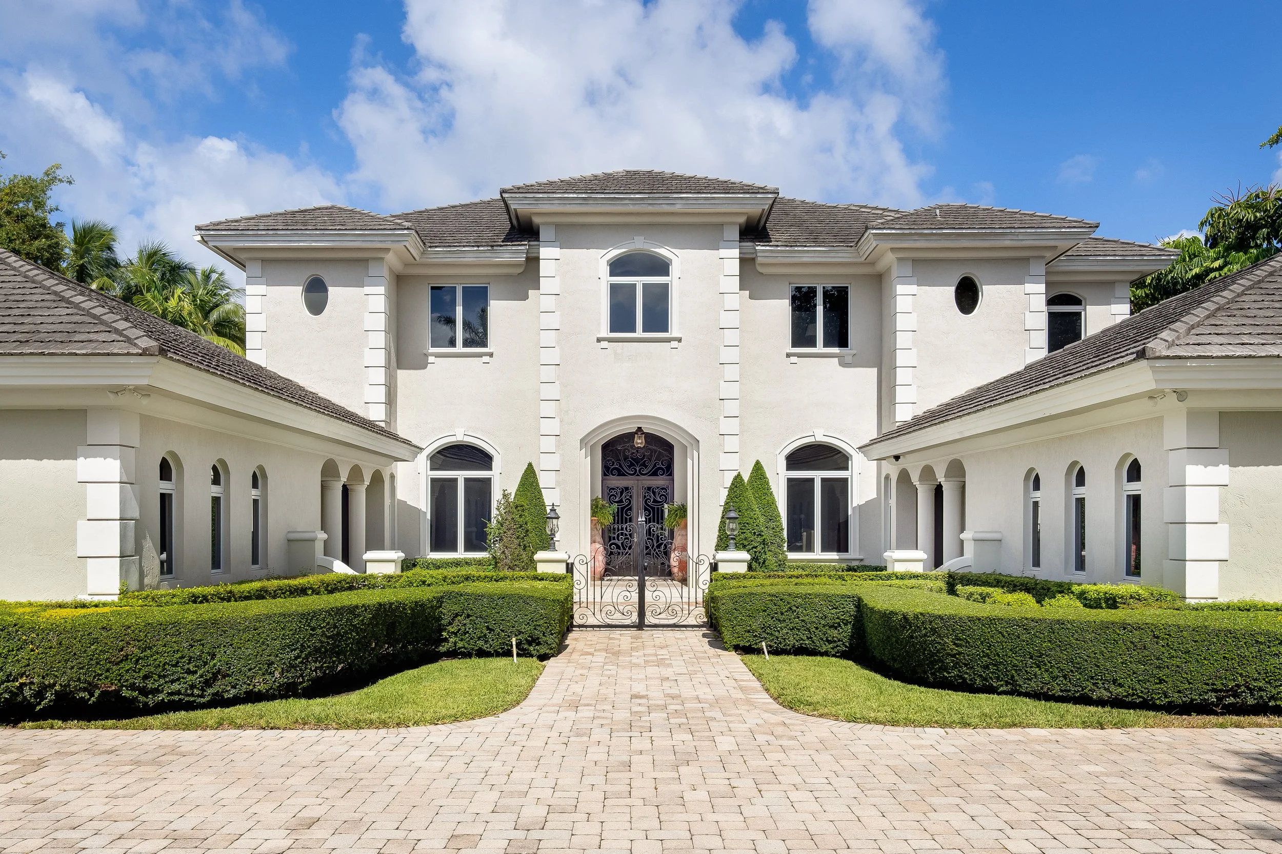 A large white house with symmetrical design, multiple windows, and a gated entrance surrounded by neatly trimmed hedges.