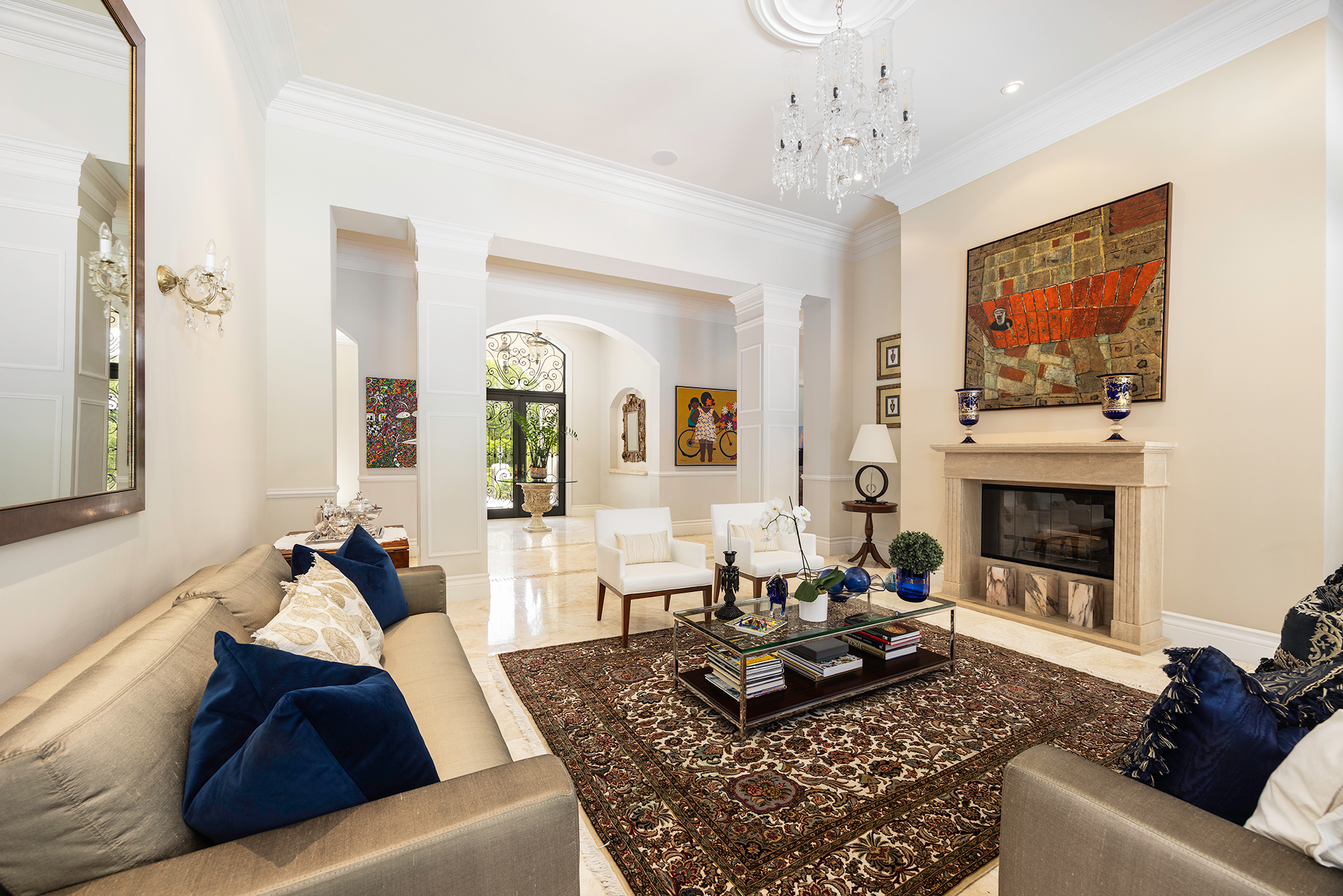 Living room with beige sofas, blue cushions, a glass coffee table with books and decorative items, a fireplace with artwork above, white chairs, a rug, and a chandelier, with an entryway in the background.
