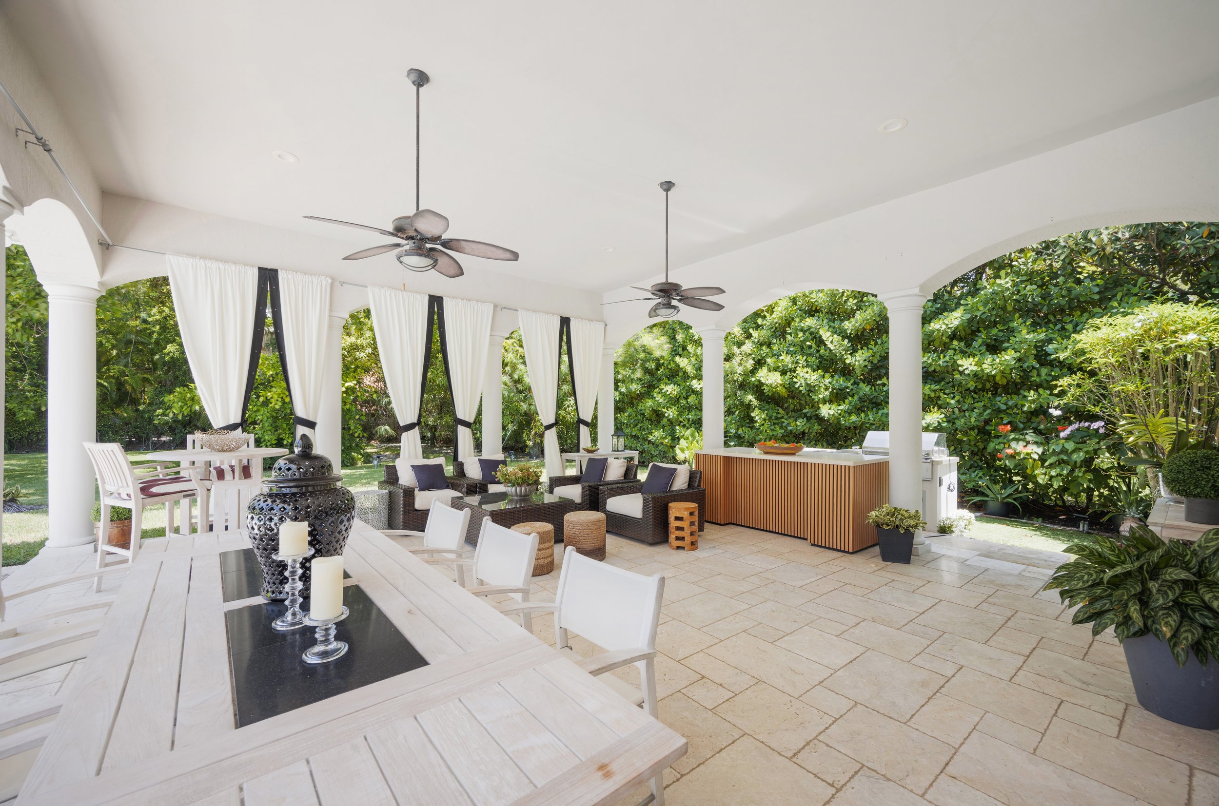 Covered outdoor patio with white curtains, ceiling fans, a dining table with candles, and a seating area with black wicker furniture and white cushions, overlooking a garden.