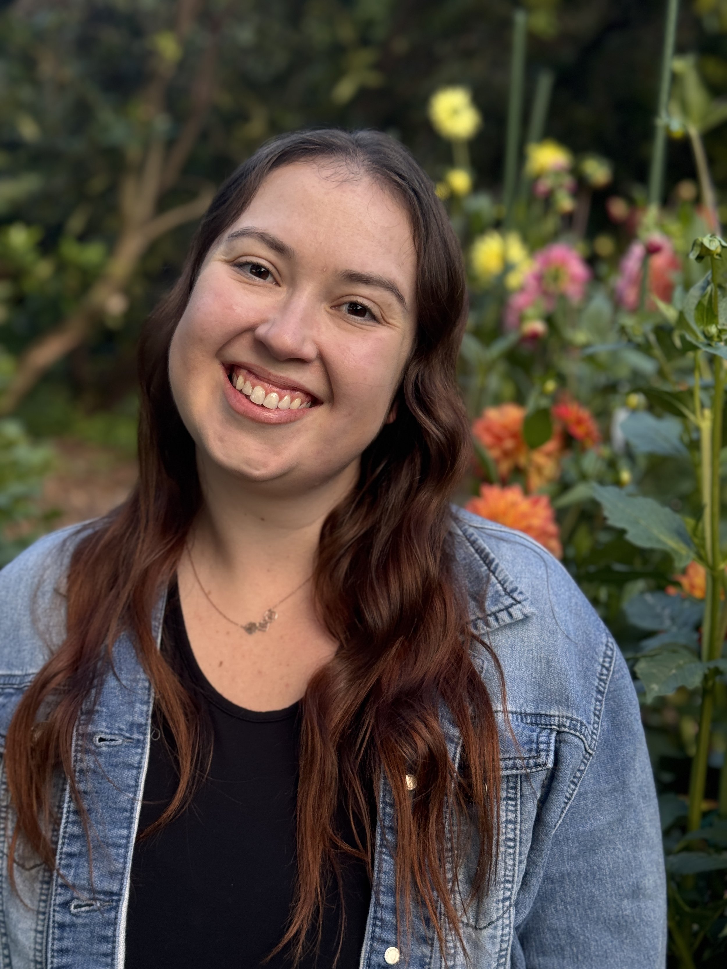 A young woman with long wavy brown hair, smiling, wearing a denim jacket and a black shirt, standing outdoors in a garden with colorful flowers and greenery in the background.