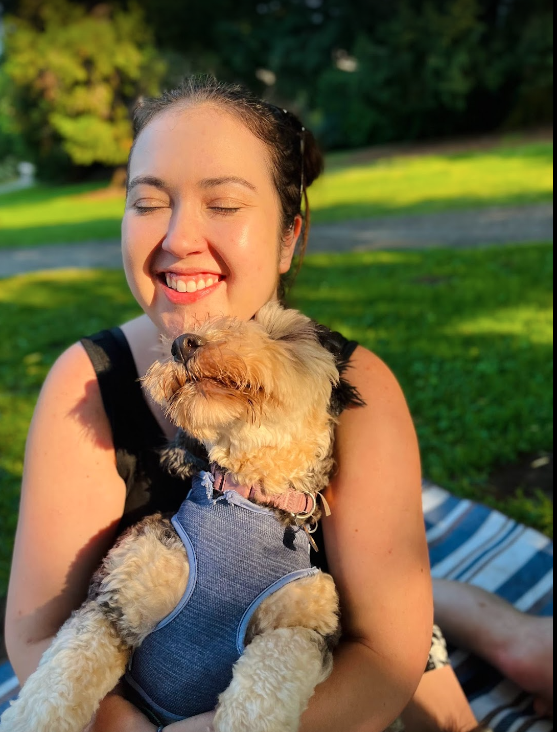 A smiling woman holding a small dog outdoors on grass, with trees in the background.