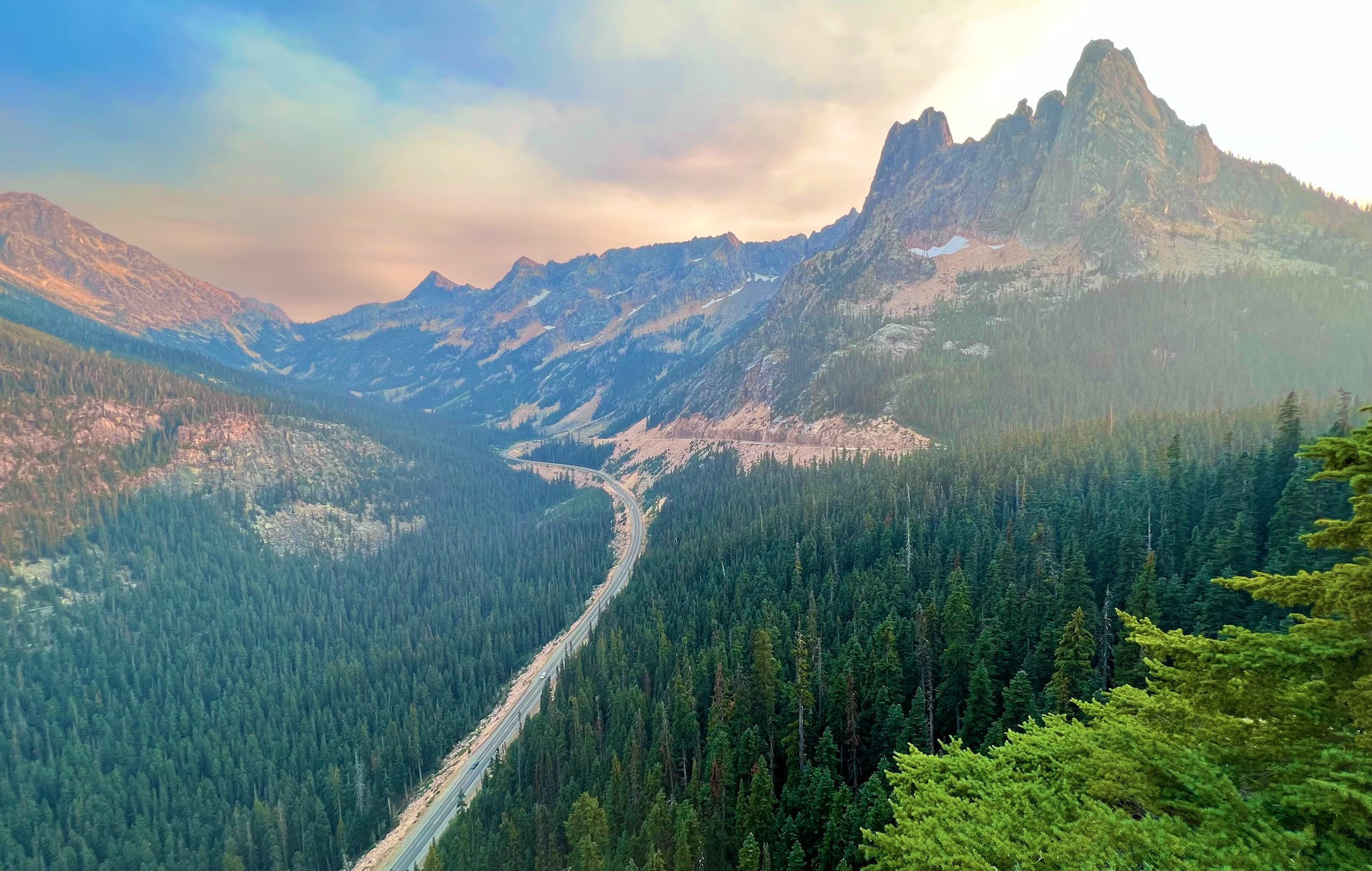 A scenic view of towering mountains with rugged peaks, a dense forest of tall green trees, and a winding highway through the valley during sunset or sunrise.