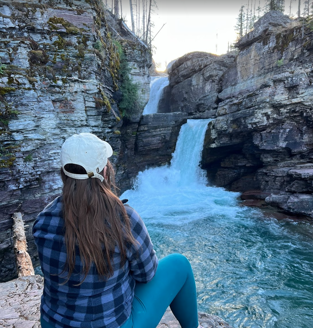 A woman with long hair wearing a white cap, blue plaid jacket, and teal pants sits on rocks, facing a waterfall in a canyon surrounded by tall rocky cliffs and trees.