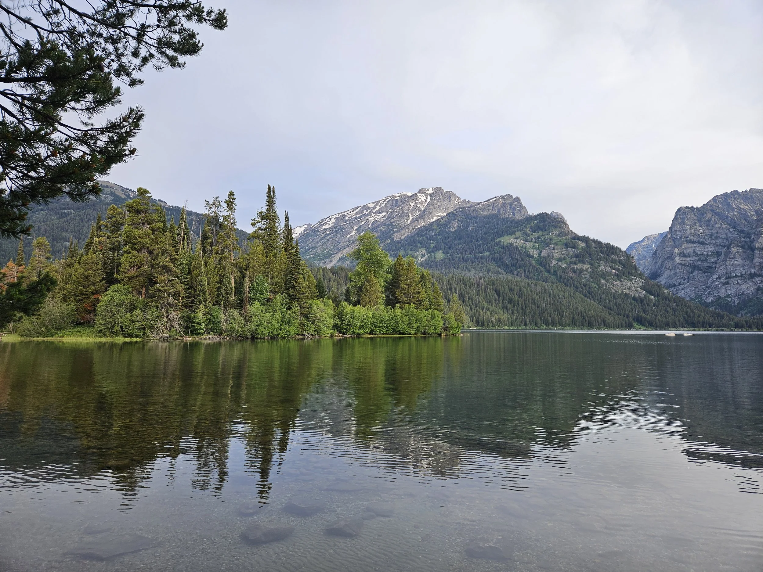 A scenic view of a lake surrounded by dense pine trees and tall mountains with patches of snow, under a partly cloudy sky.