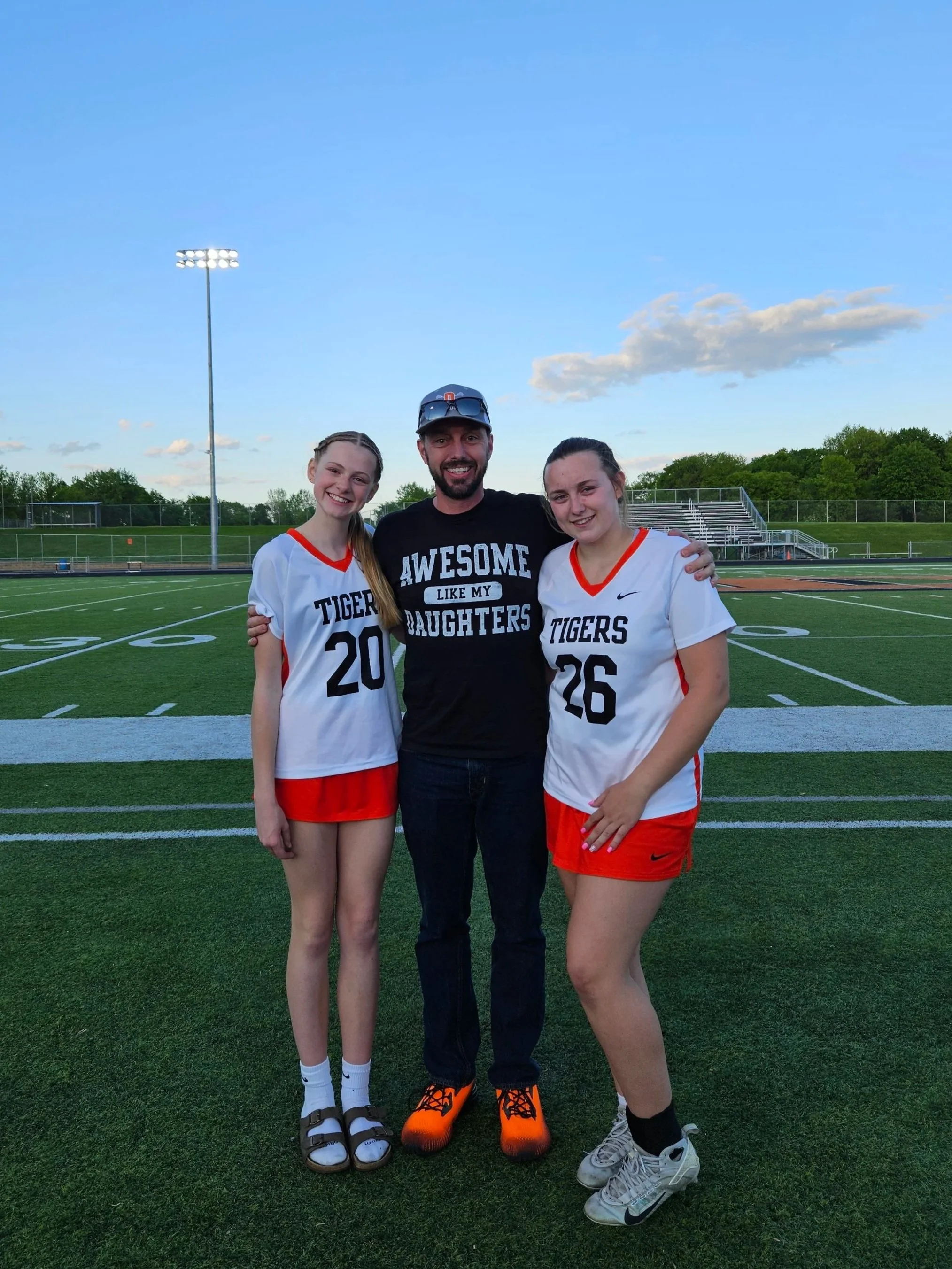 Two young female athletes in white soccer jerseys with red accents and a man in a black T-shirt with white text, standing on a sports field. The women have their arms around the man, and they are smiling at the camera during the daytime.