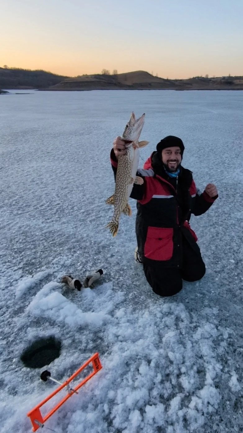 A man kneeling on a frozen lake holding a fish he caught, with ice fishing gear and a hole in the ice nearby, at sunset or sunrise.
