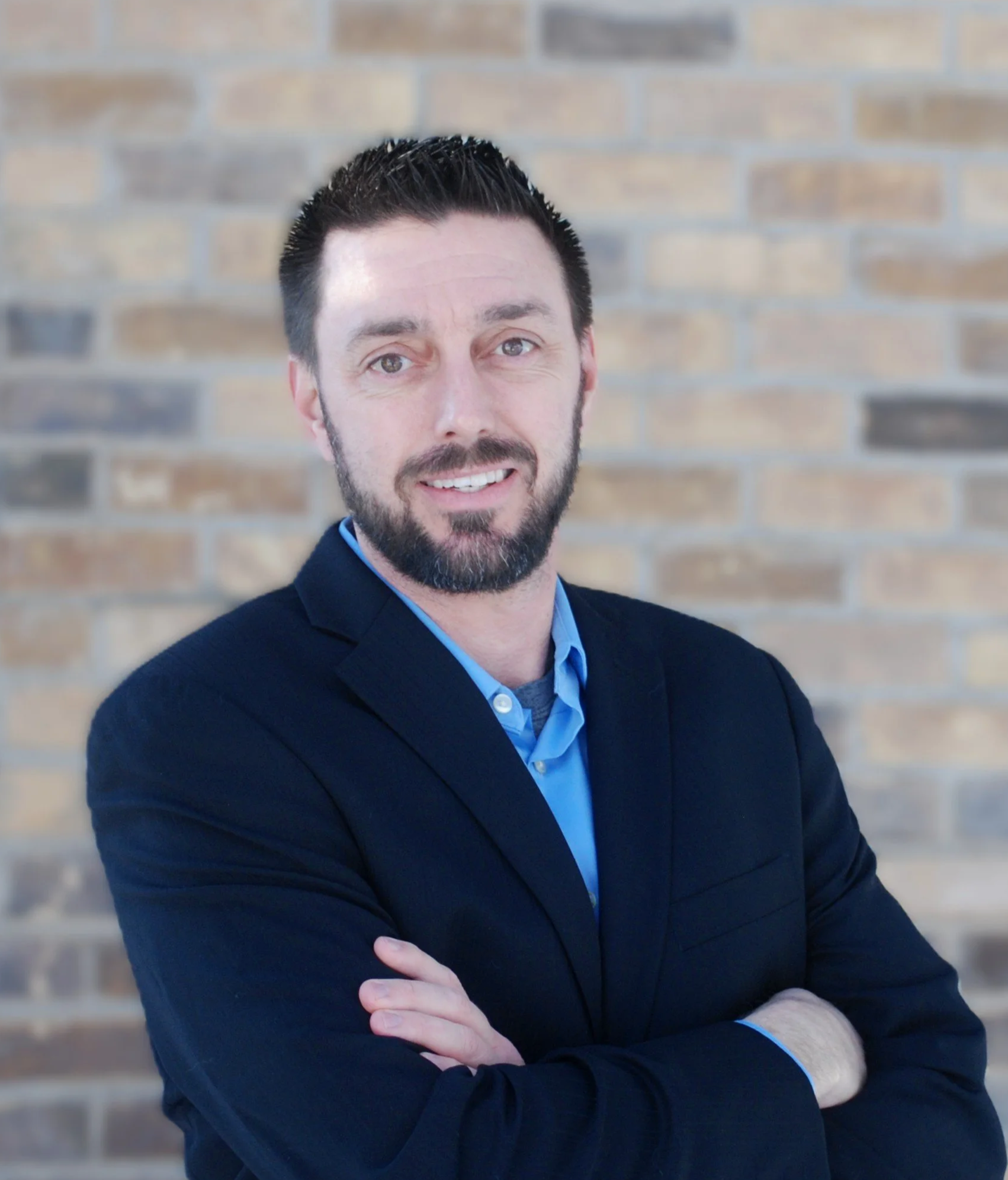 A man with dark hair and a beard, smiling and wearing a dark blazer and blue shirt, standing in front of a brick wall.