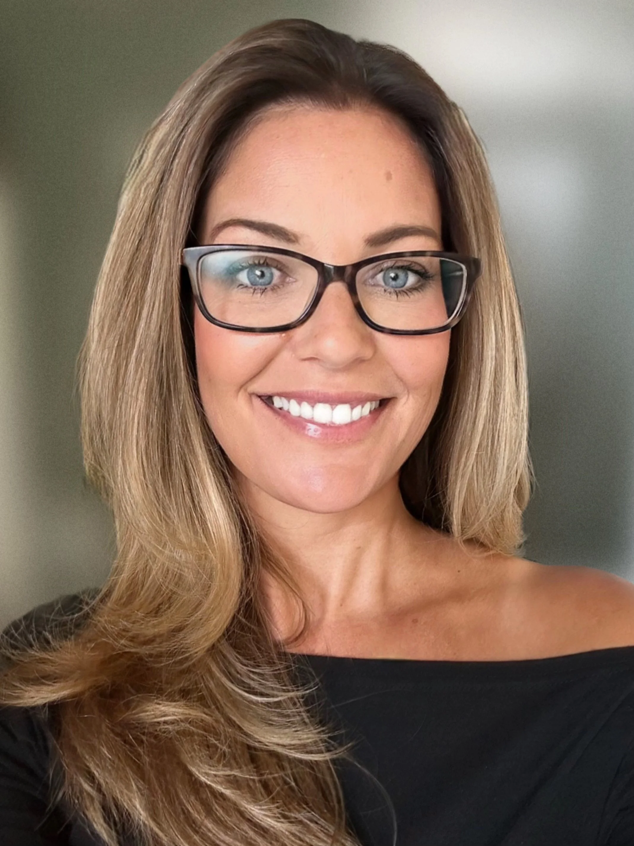 A smiling woman with long, wavy, light brown hair and glasses, wearing a black off-the-shoulder top, against a neutral background.