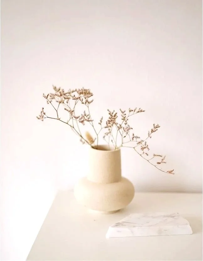 A cream-colored vase with a rounded bottom and a tall, cylindrical neck holding dried delicate white flowers, placed on a white surface next to a small white marble slab, against a plain light background.