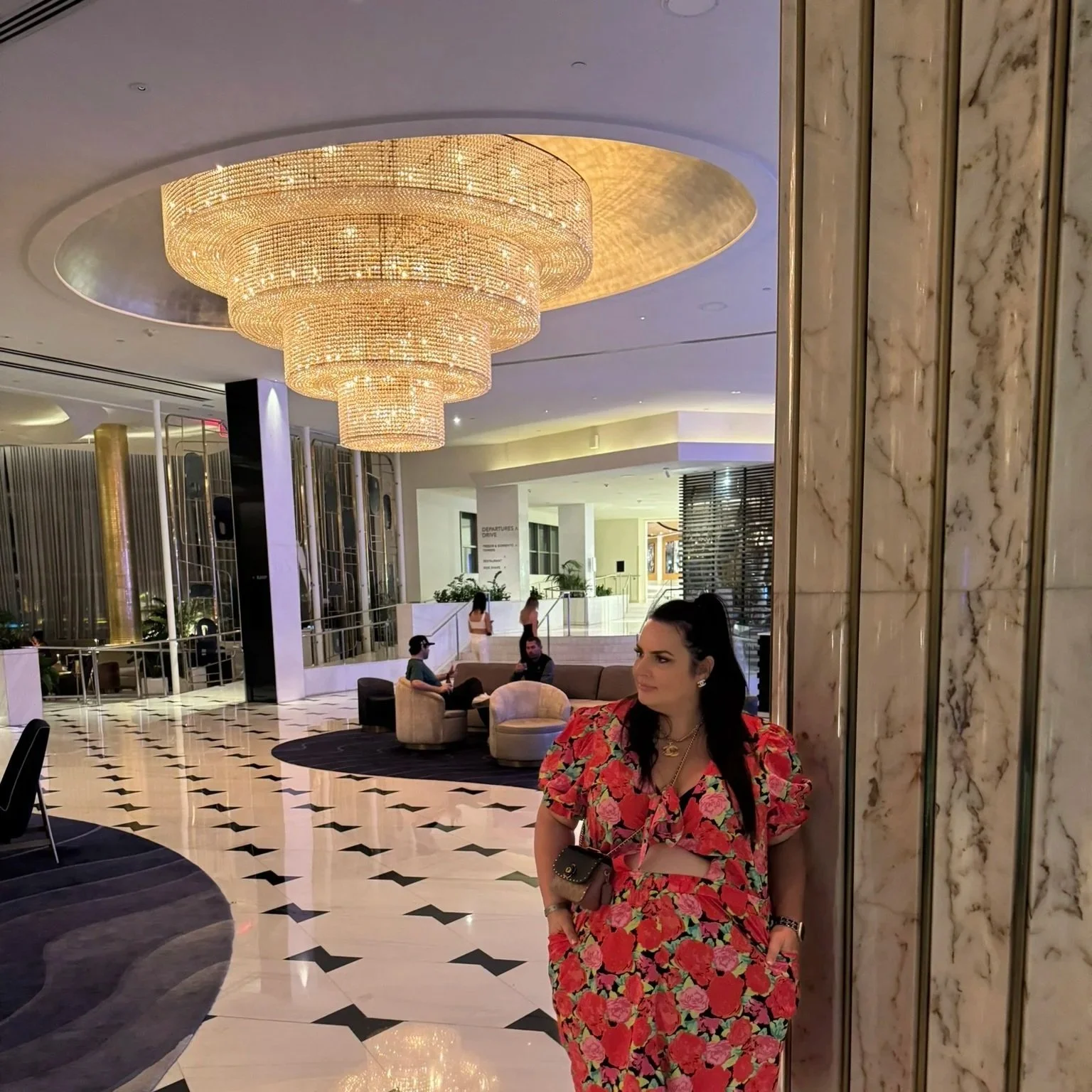 A woman in a red floral dress leaning against a marble pillar in a Fontaine Bleau Miami hotel lobby with a large crystal chandelier overhead and seating area in the background.