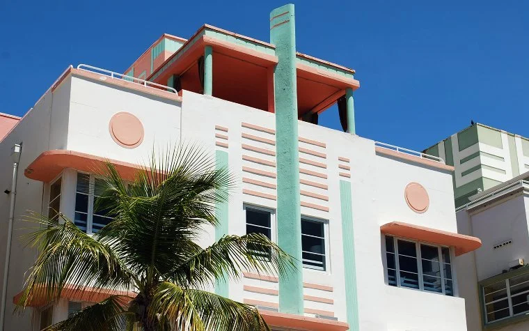 Art Deco style building with pastel colors, rounded features, and tall vertical elements, with a clear blue sky in the background and a palm tree in front.