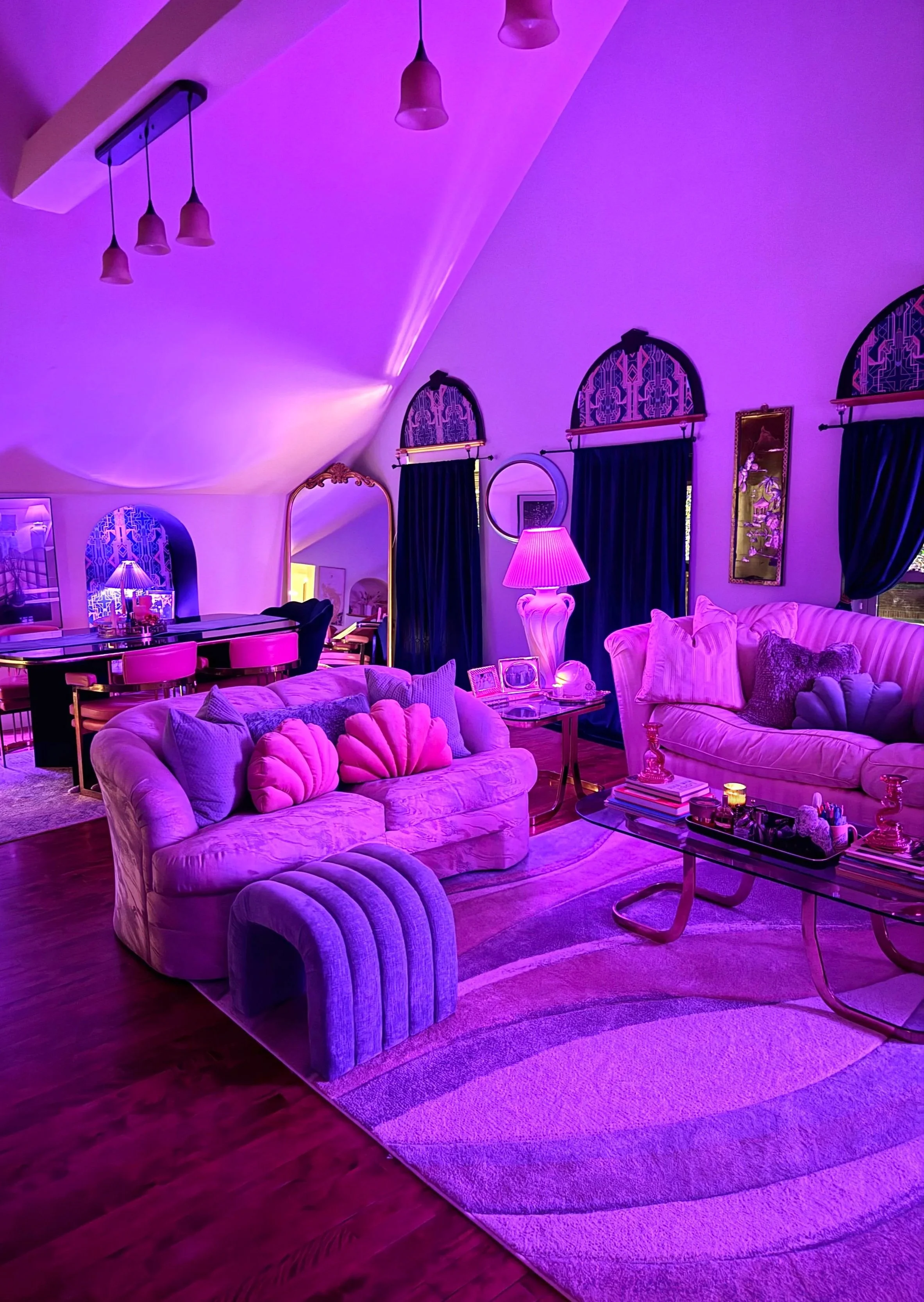 A living room illuminated with purple lighting, featuring plush sofas with decorative pillows, a glass coffee table with books and candles, a tall pink lamp, dark curtains, and windows with ornate valances.