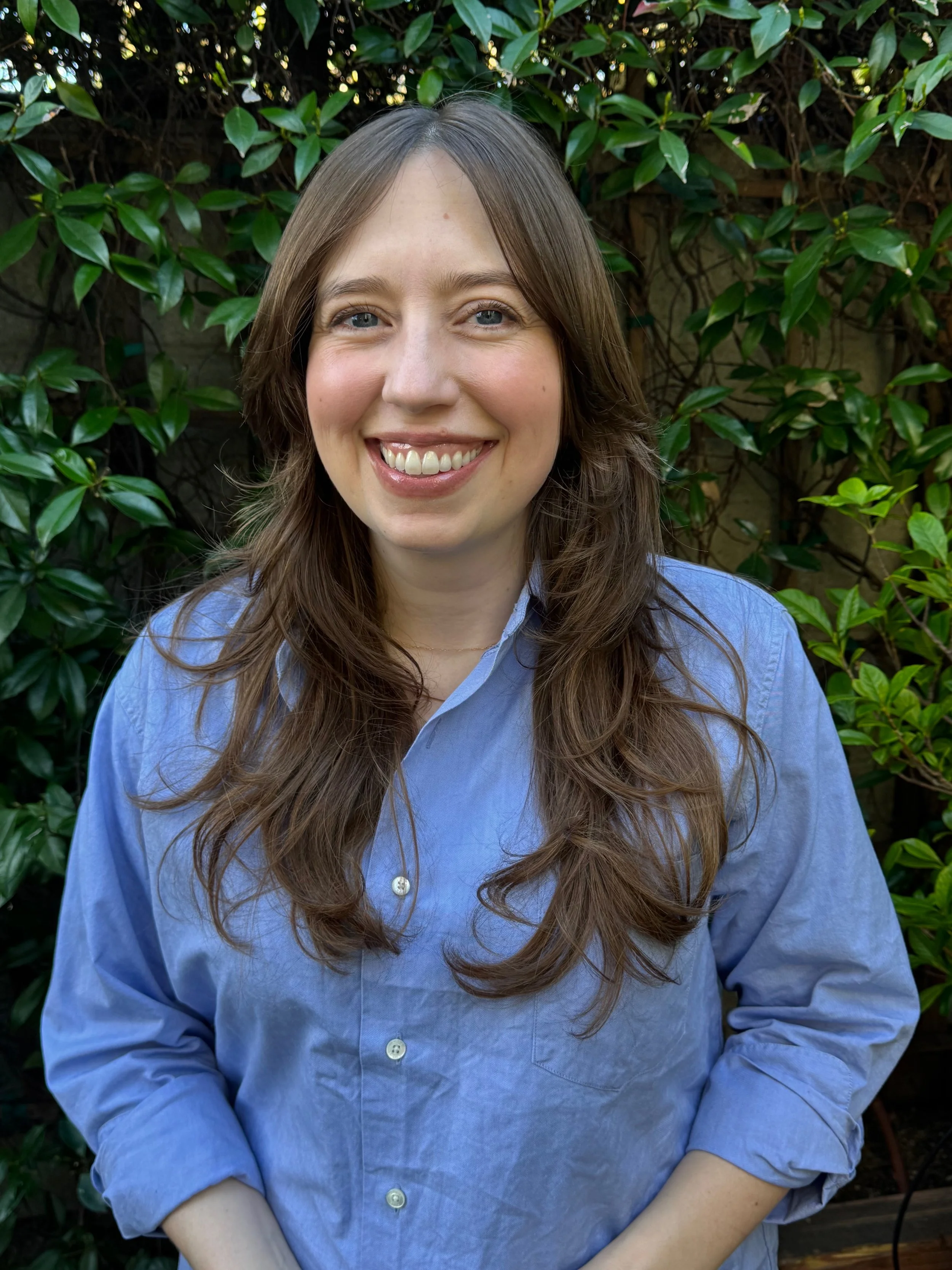 A woman with long brown hair wearing a light blue button-down shirt standing outdoors in front of green leafy plants, smiling at the camera.