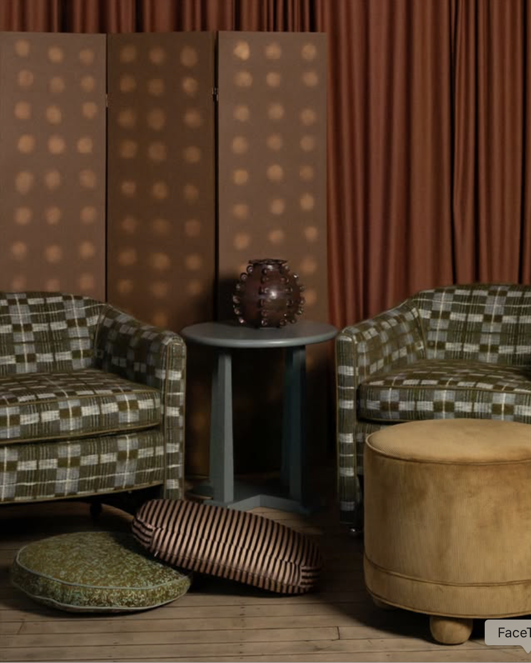 A living room setup with two patterned sofas, a small side table with a decorative vase, behind which are orange curtains and a patterned folding screen. There are various cushions and pillows on the wooden floor in front.