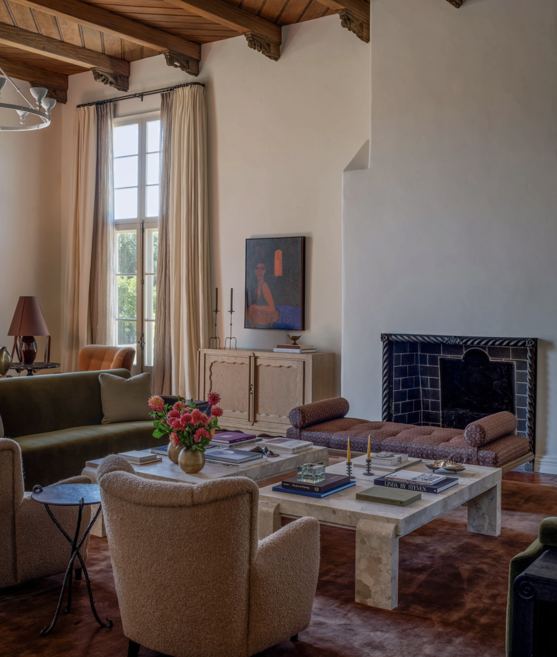 Living room with high vaulted wooden ceiling, large window with beige curtains, beige walls, a stone fireplace, and a variety of furniture including a green sofa, beige armchair, coffee table with books and flowers, and a credenza with artwork and decorative items.
