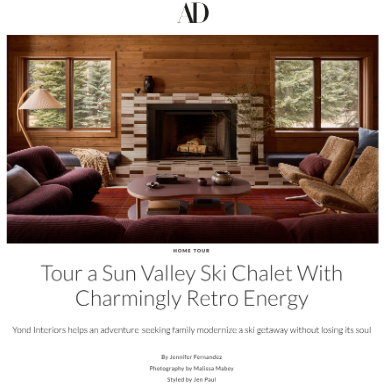 Living room with wood-paneled walls, a fireplace with a patterned tile surround, two large windows showing outdoor trees, and cozy seating including armchairs and a sofa around a central coffee table.