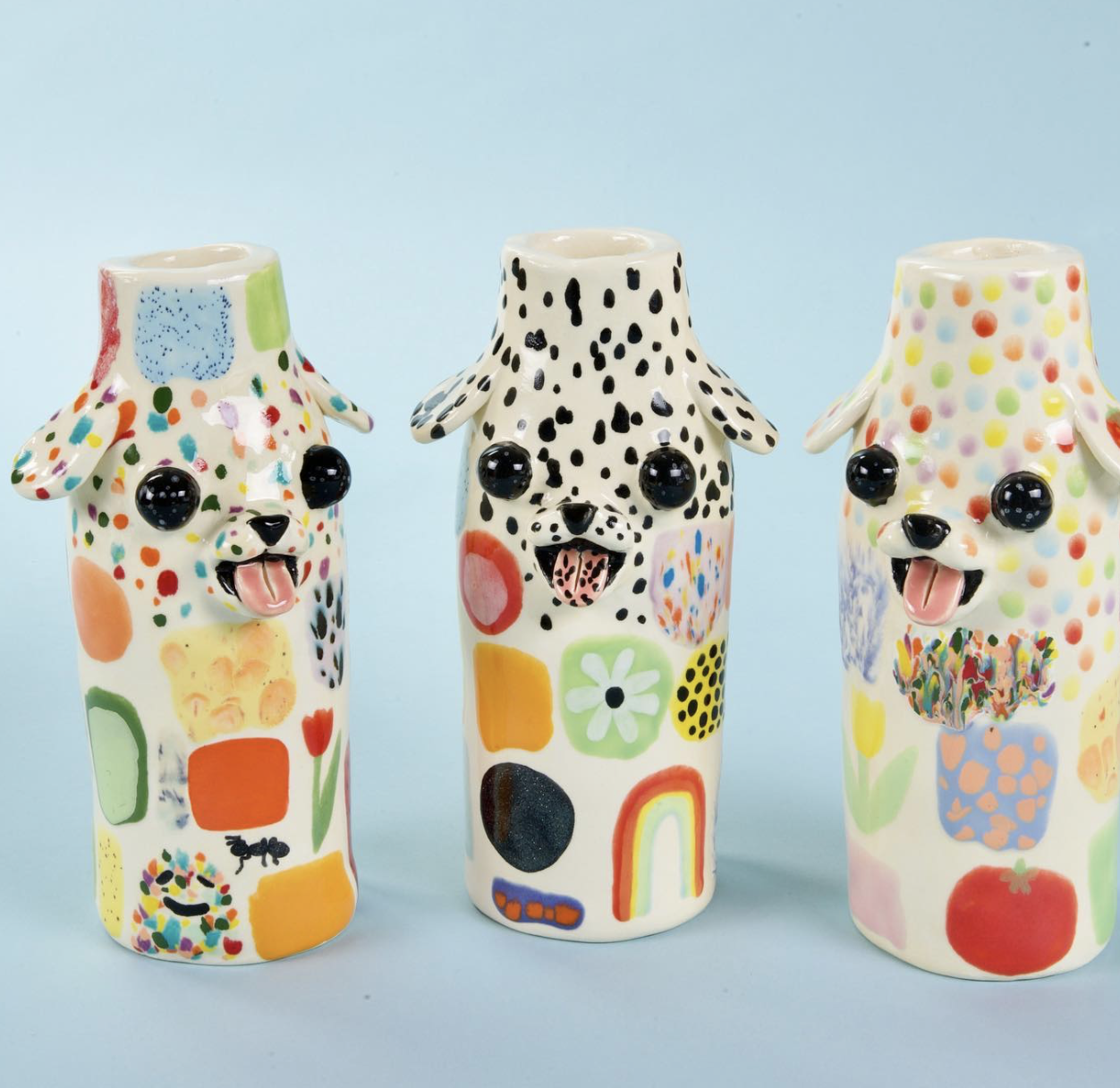 Three colorful ceramic dog-shaped vases with painted floral and abstract designs, each with a different pattern of dots and shapes, and black glossy eyes and open mouths with tongues, set against a light blue background.