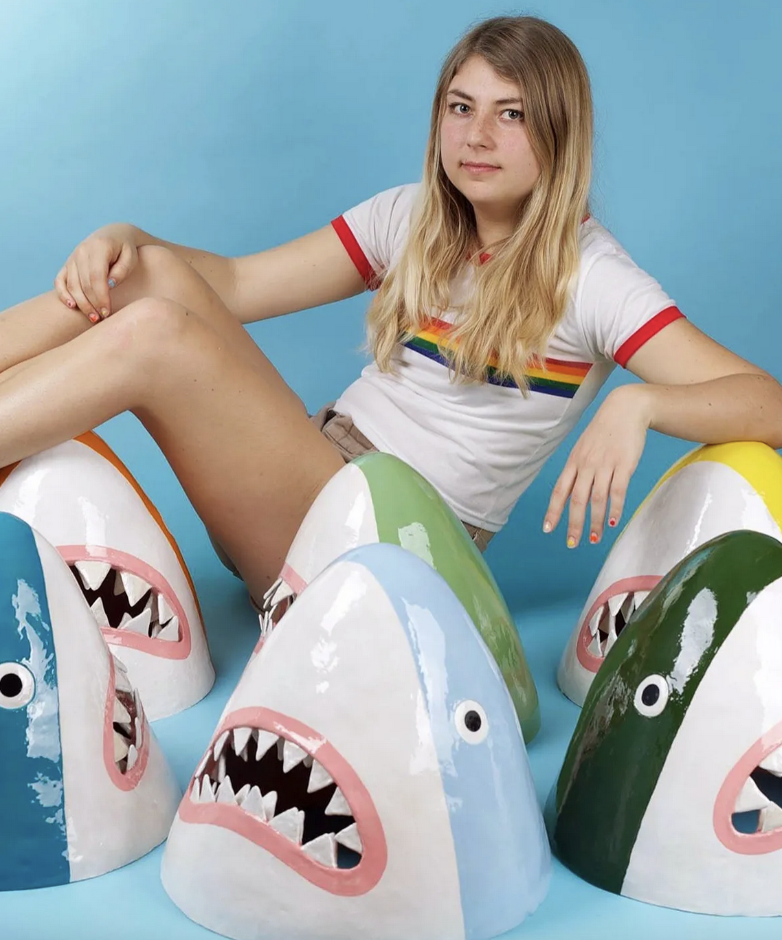A young woman with long blonde hair sitting on colorful shark-shaped objects, wearing a white t-shirt with a rainbow stripe, against a blue background.